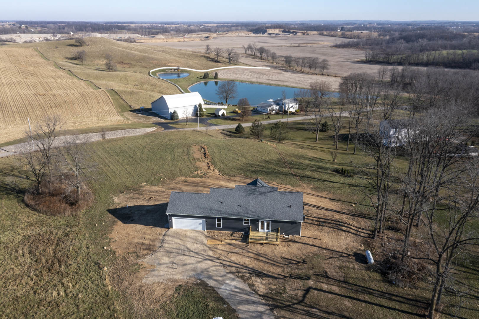 White house with gabled roof set in grassy field, surrounded by mature trees and a small pond, wooden deck visible in foreground, rural landscape under open sky