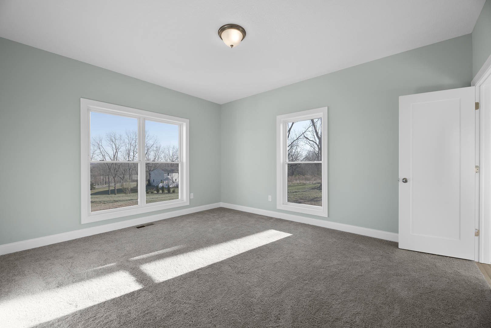 Carpeted room with white walls, white door with silver knob, two windows showing trees and field, ceiling light fixture, crown molding