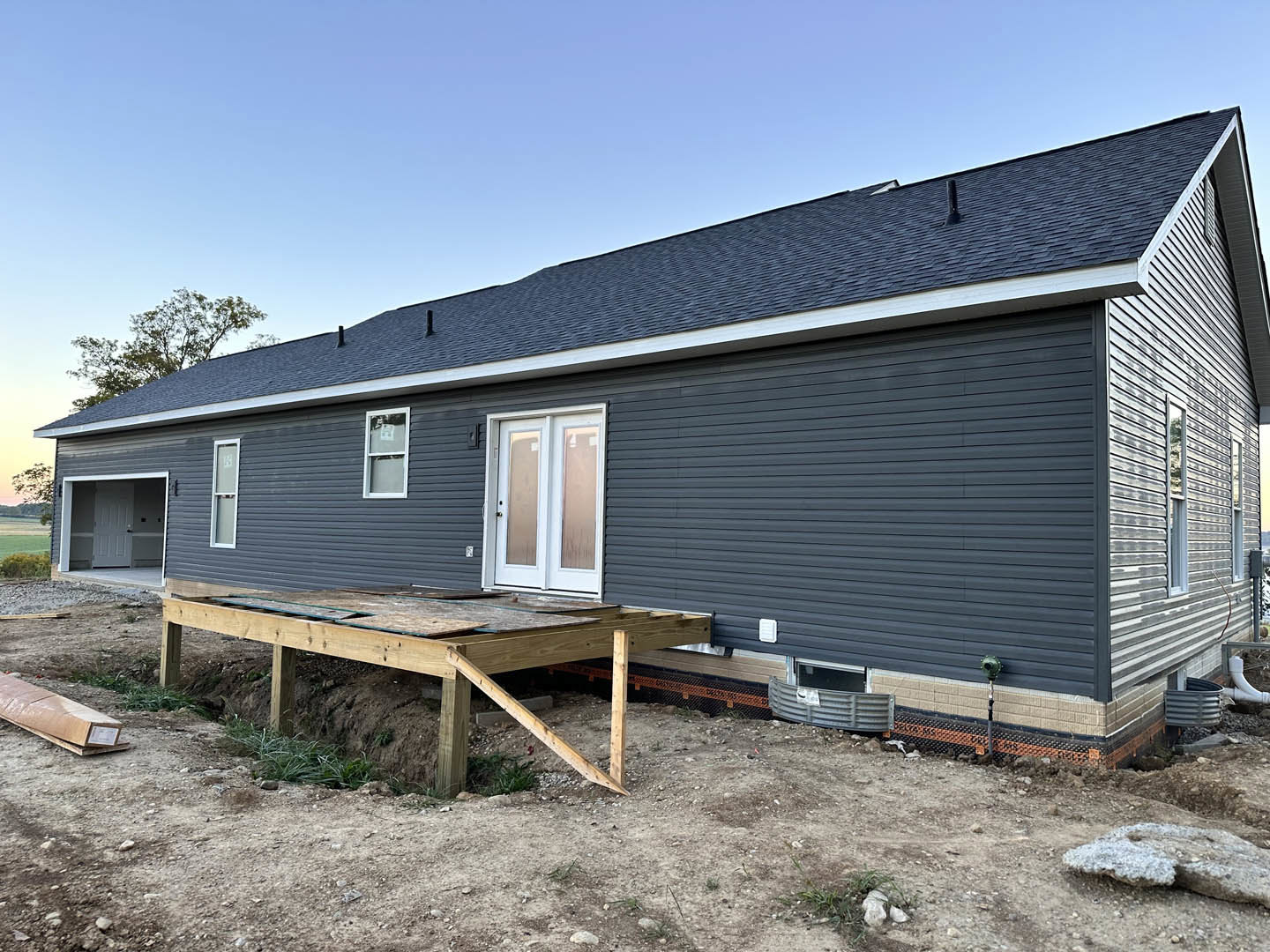 Wooden deck with natural finish extends from house featuring white siding, glass-paneled white door, and white-framed window; porch area partially covered, cardboard piece rests on
