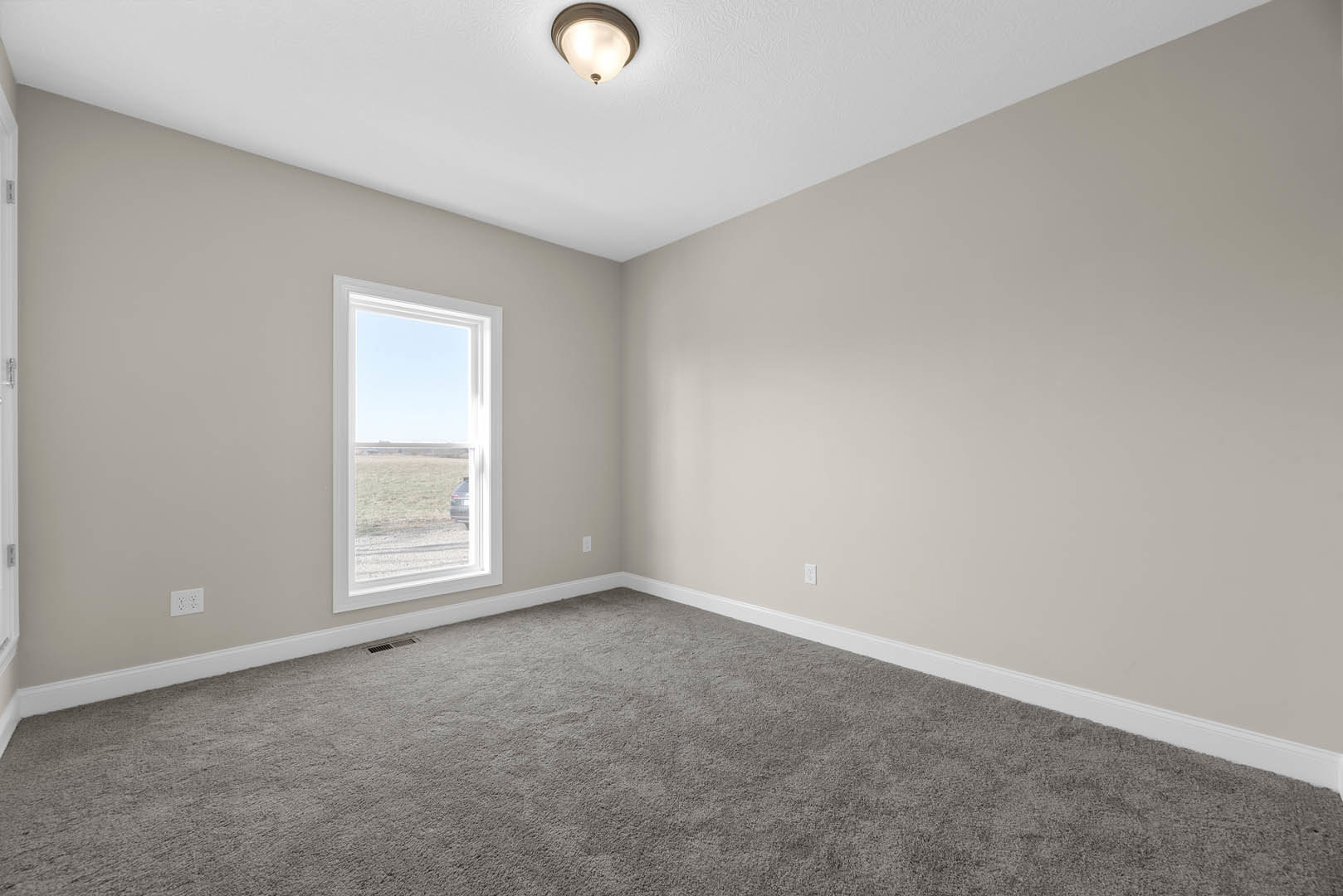 Bright room with grey carpet, white walls, large window showing blue sky and parked car, ceiling-mounted light fixture, simple molding along ceiling and floor.