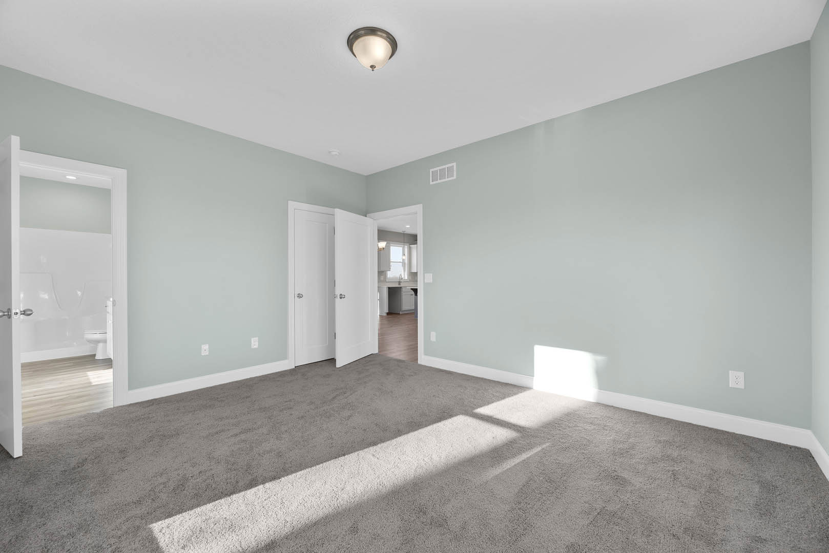 Light blue walls and carpeted floor, ceiling light fixture, white bathroom with toilet and tub visible, white door with silver knob, light shining onto carpet.