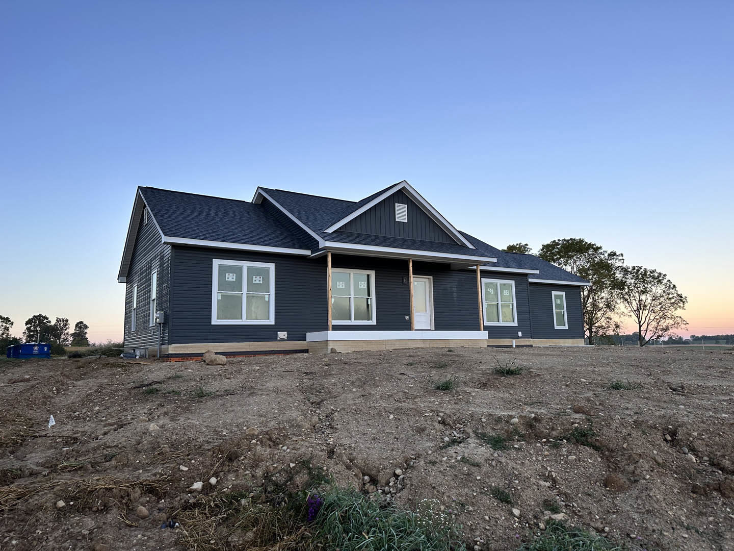 Partially built house with exposed framing, blue roof, white window frames, covered porch, large excavation in front yard, surrounded by trees under clear blue sky