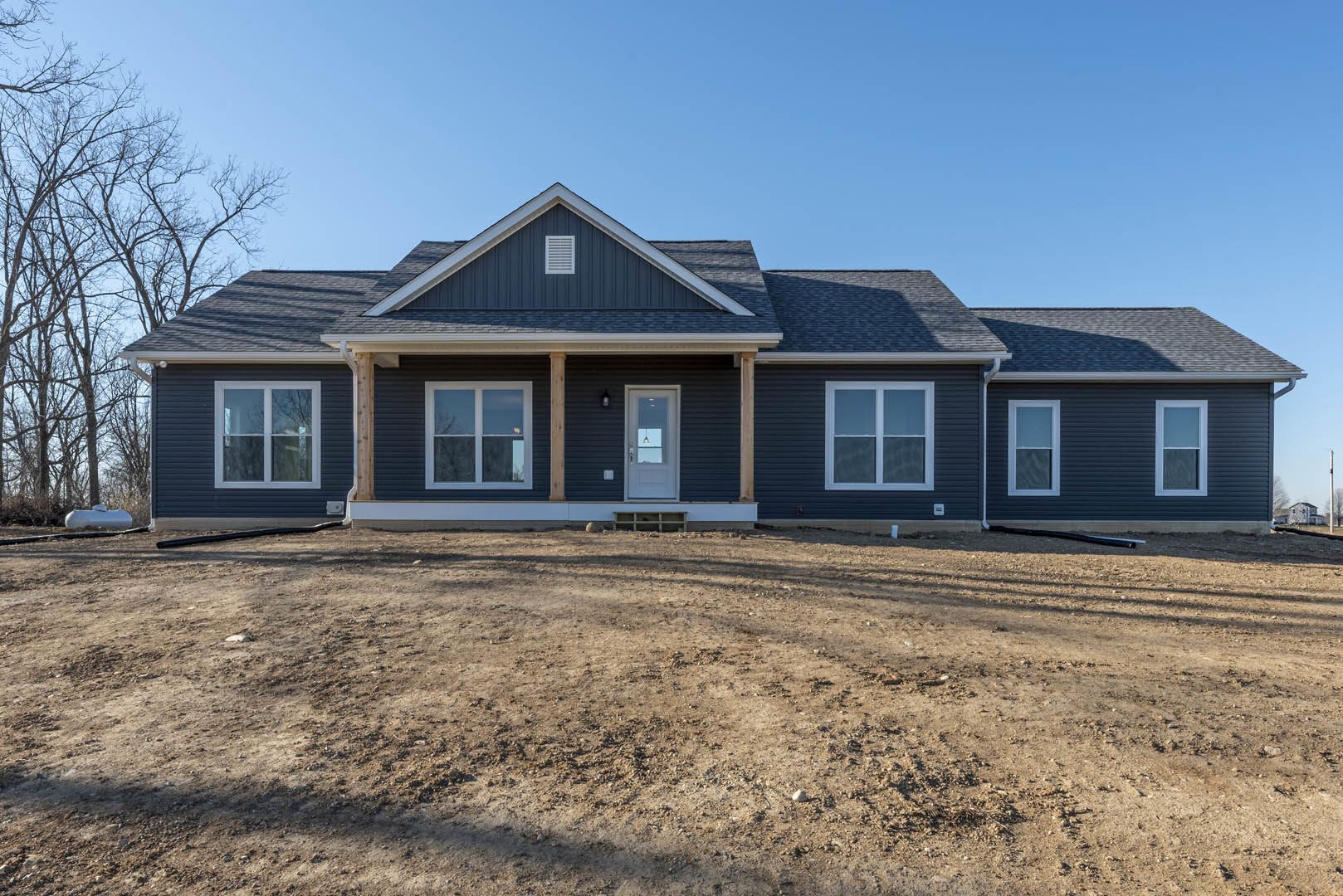 Two-story house under construction with exposed framing, white window frames, dirt yard, and surrounding trees