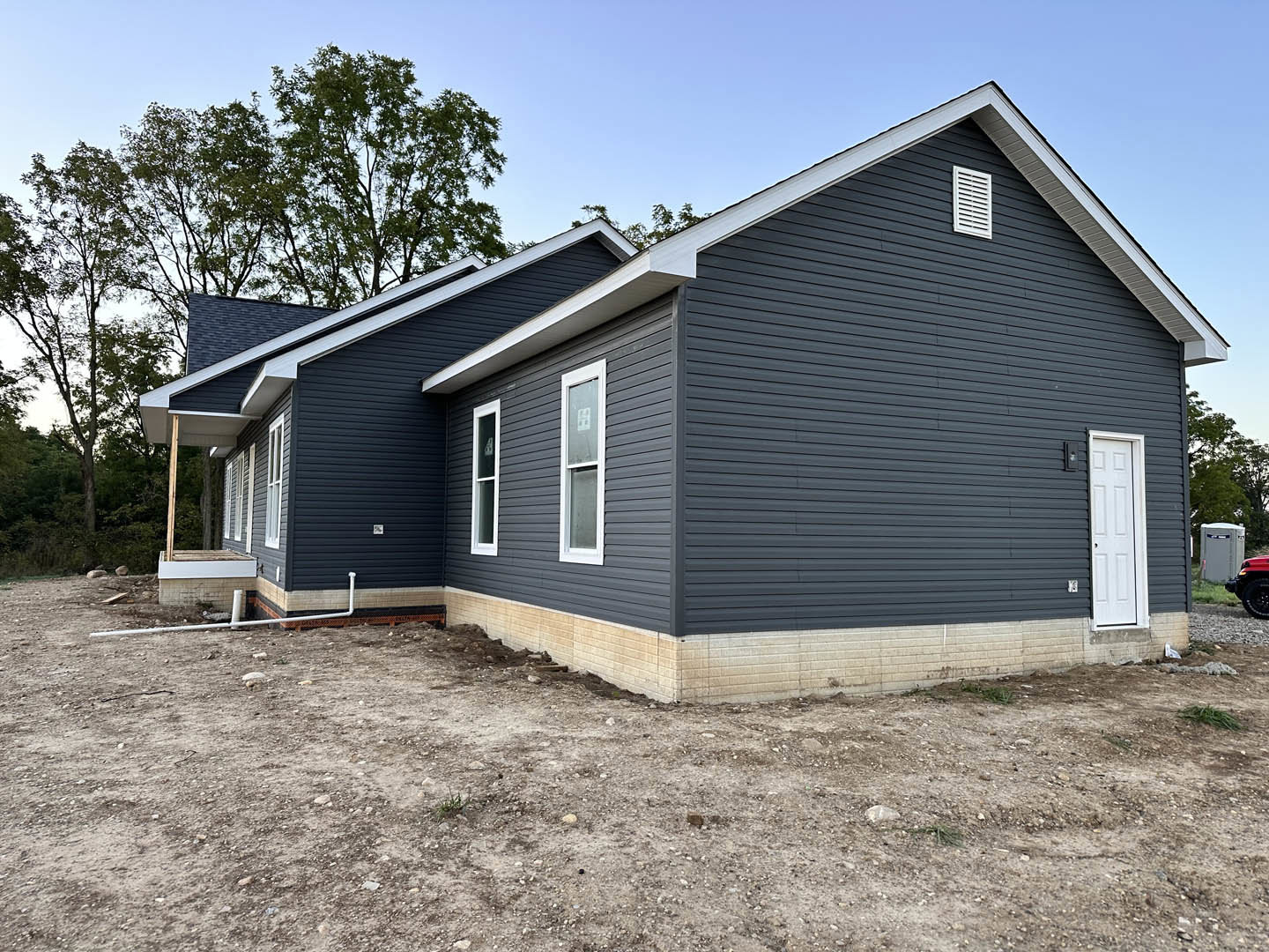 Modern home with black siding, white door, and white-framed window; dirt yard in front, tree in background, grey portable toilet on roadside.