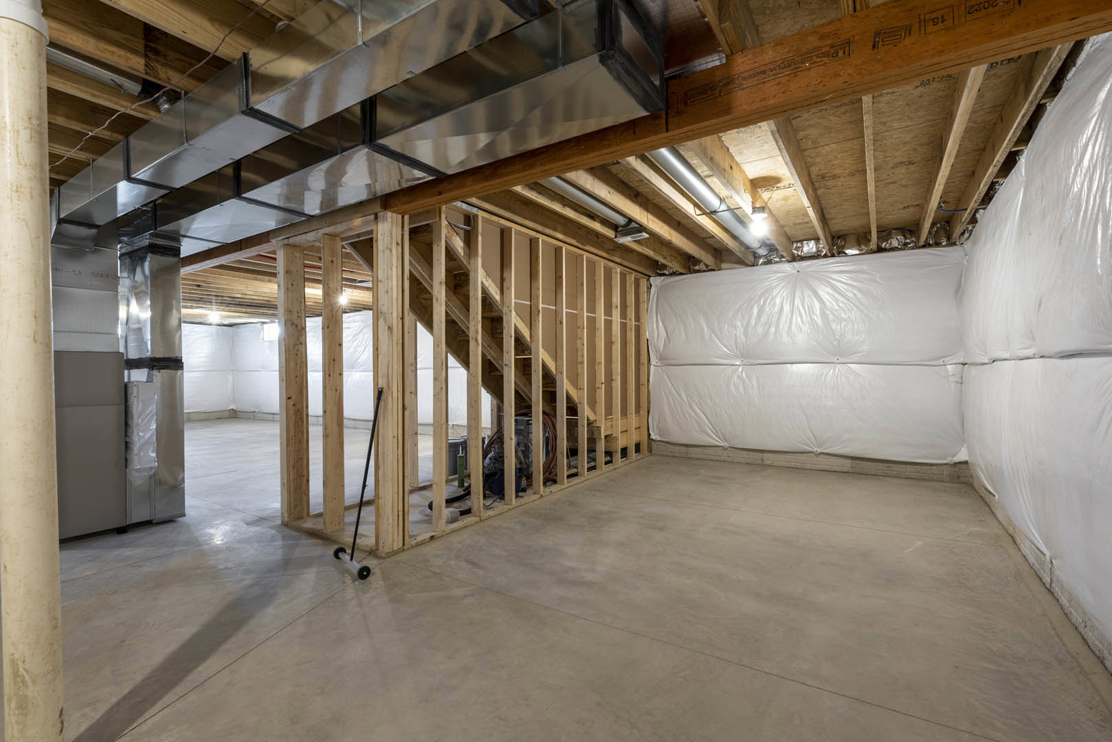 Open-concept room with exposed wood framing, concrete floor, white plaster wall, staircase, steel beam, and plastic insulation covering