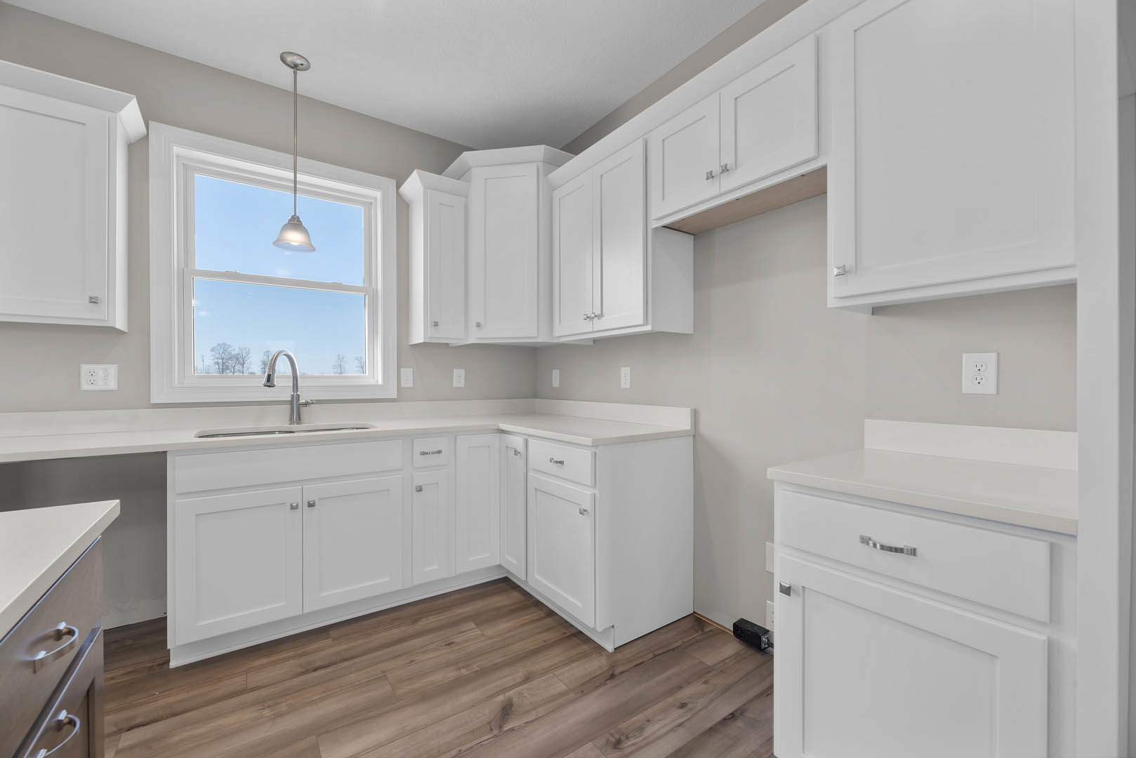 White kitchen cabinets with brushed metal handles, wood flooring, a stainless steel sink and faucet beneath a window, white electrical outlet on the wall, and a ceiling-mounted