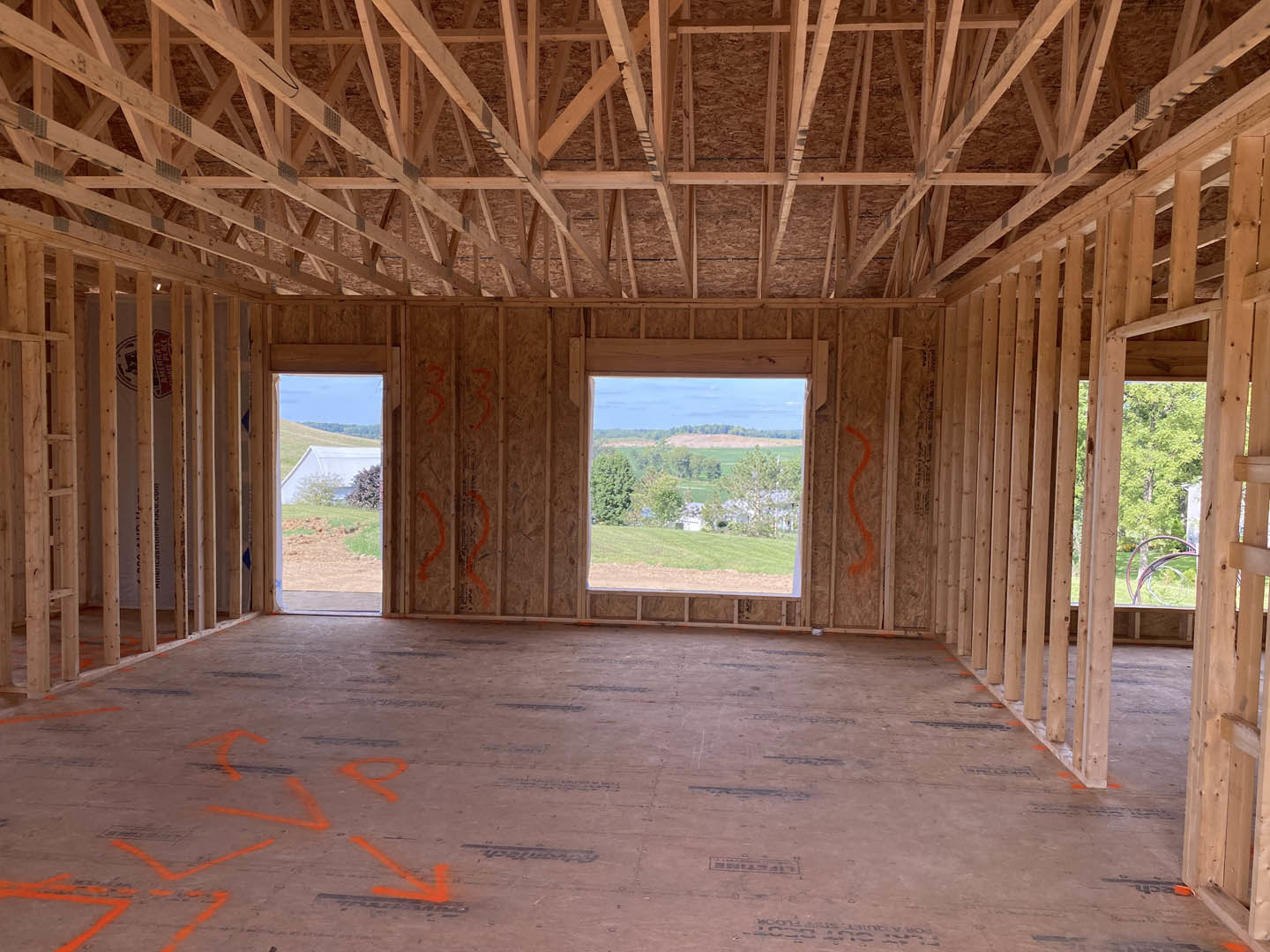 Open-concept room with exposed wood beam ceiling, large window overlooking grassy field and trees, light-toned flooring, and natural daylight streaming in