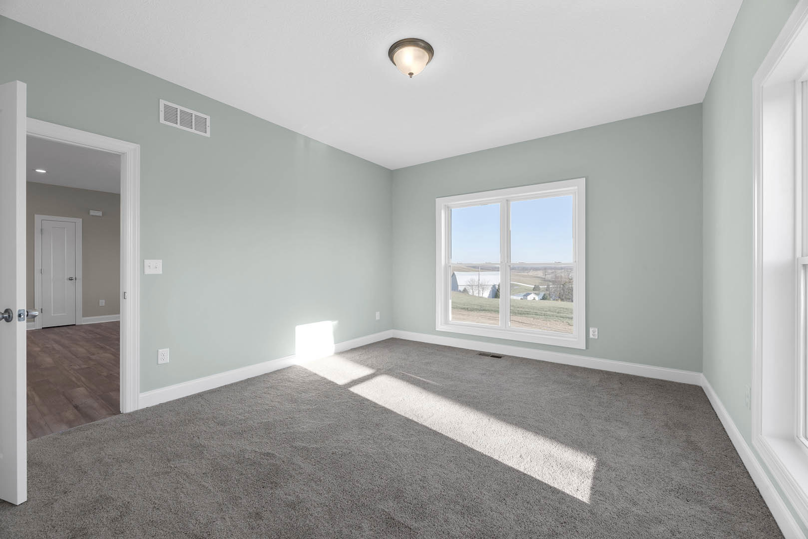 Carpeted room with white walls, large window showing farm view, white door with silver handles, ceiling light fixture, wall vent, sunlight streaming through window