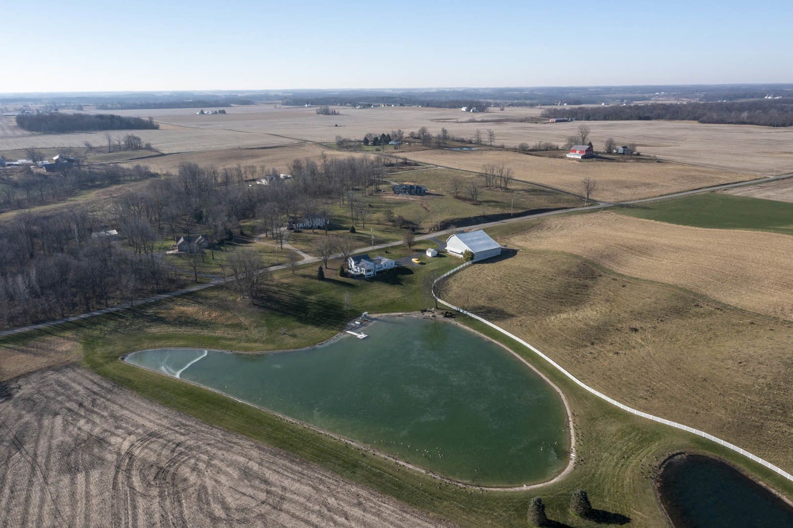 White house with dark roof and several windows beside grassy field and pond with ducks and small boat, blue sky overhead, scattered trees in background.