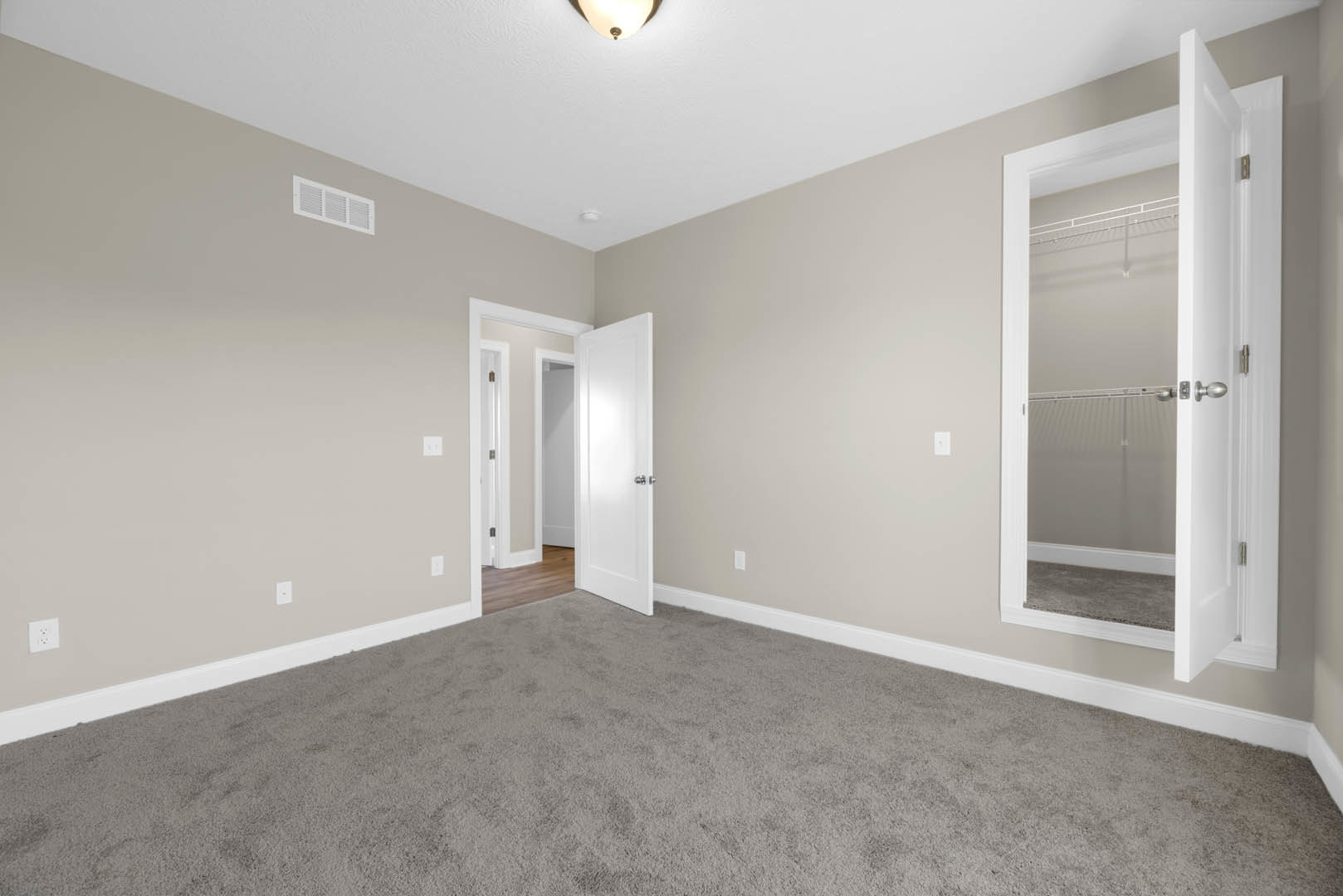 Bedroom with beige carpet, open white closet door, wall-mounted mirror, silver door hardware, white ceiling vent, and light-colored walls with crown molding