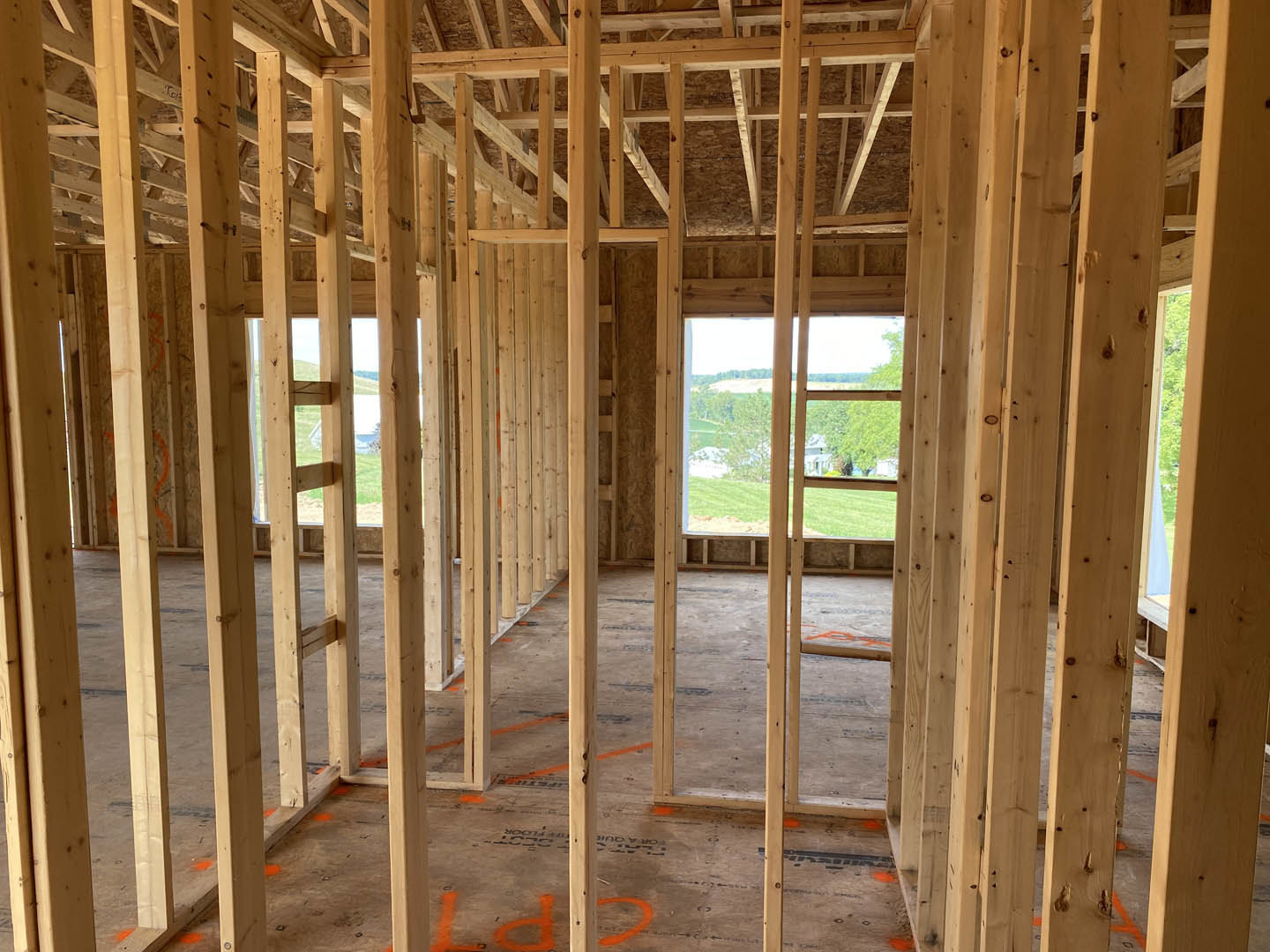 Exposed wood framing and beams inside a partially constructed home, window opening reveals trees and grassy field outside