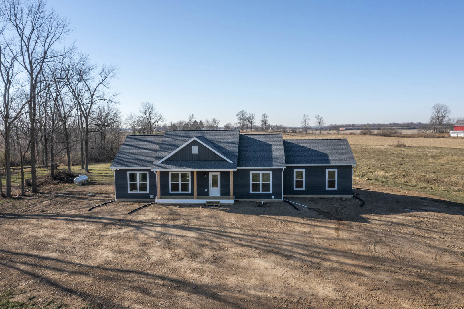 Partially built house with white front door, covered porch, and unfinished dirt yard, surrounded by grassy field and scattered trees under blue sky with clouds