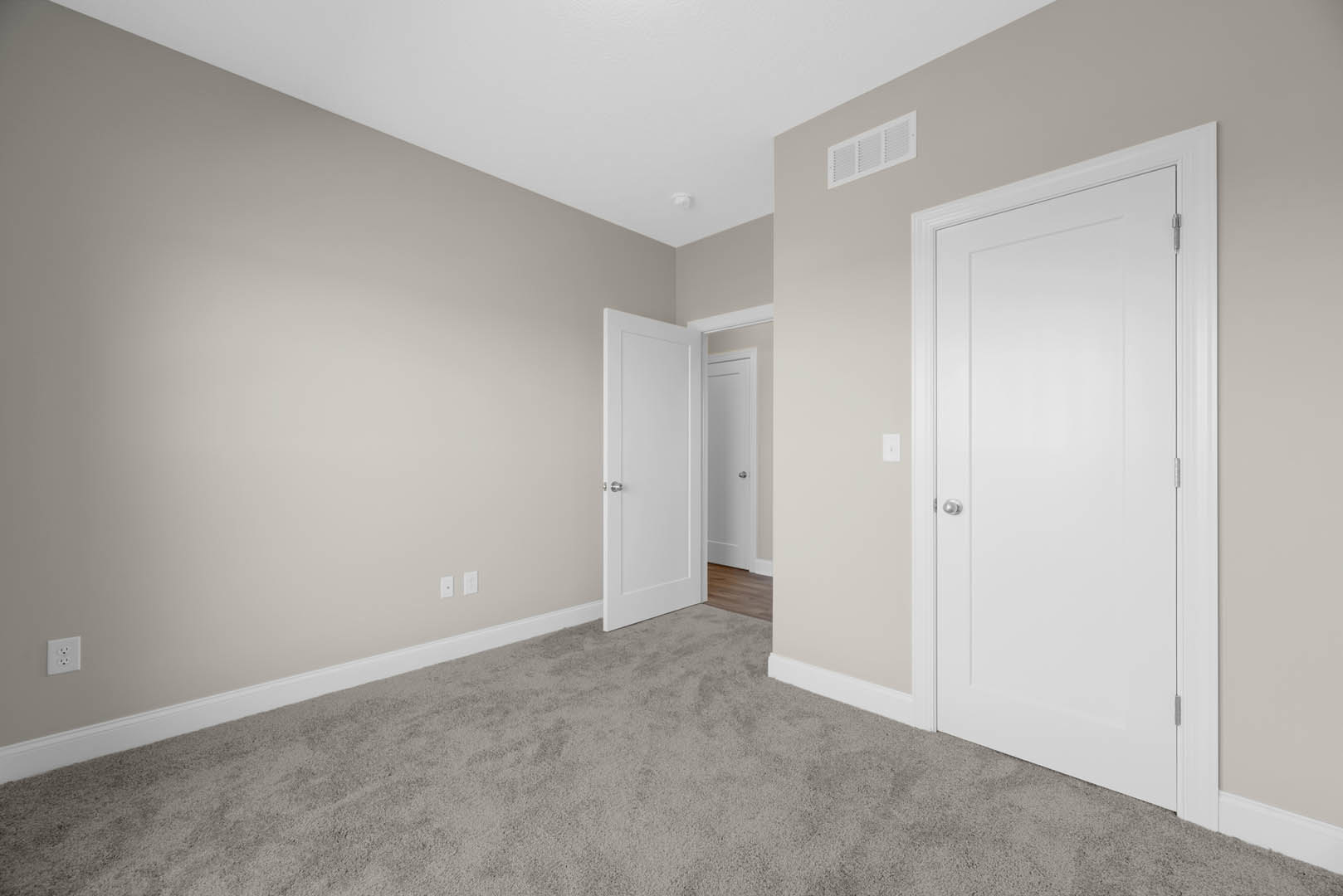 White carpeted room featuring multiple white paneled doors with silver knobs, white electrical outlet and wall vent, light-colored walls with base molding.