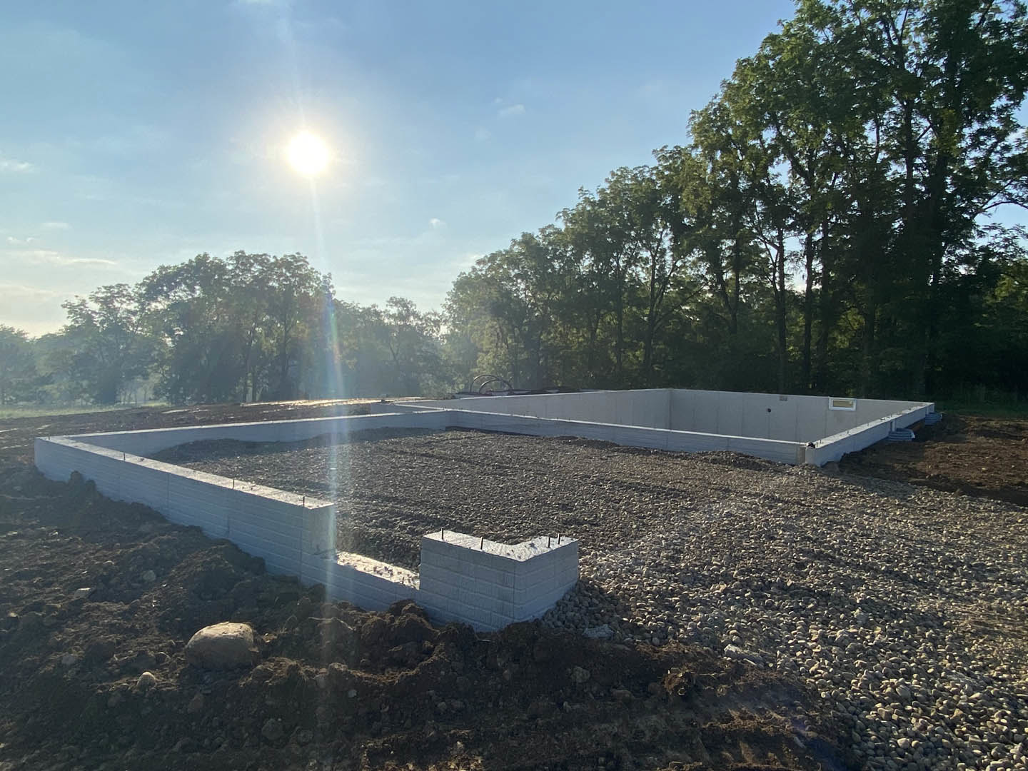 Concrete foundation wall with exposed rebar, pile of dirt nearby, large rock in foreground, bright sunlight filtering through surrounding trees, clear sky overhead