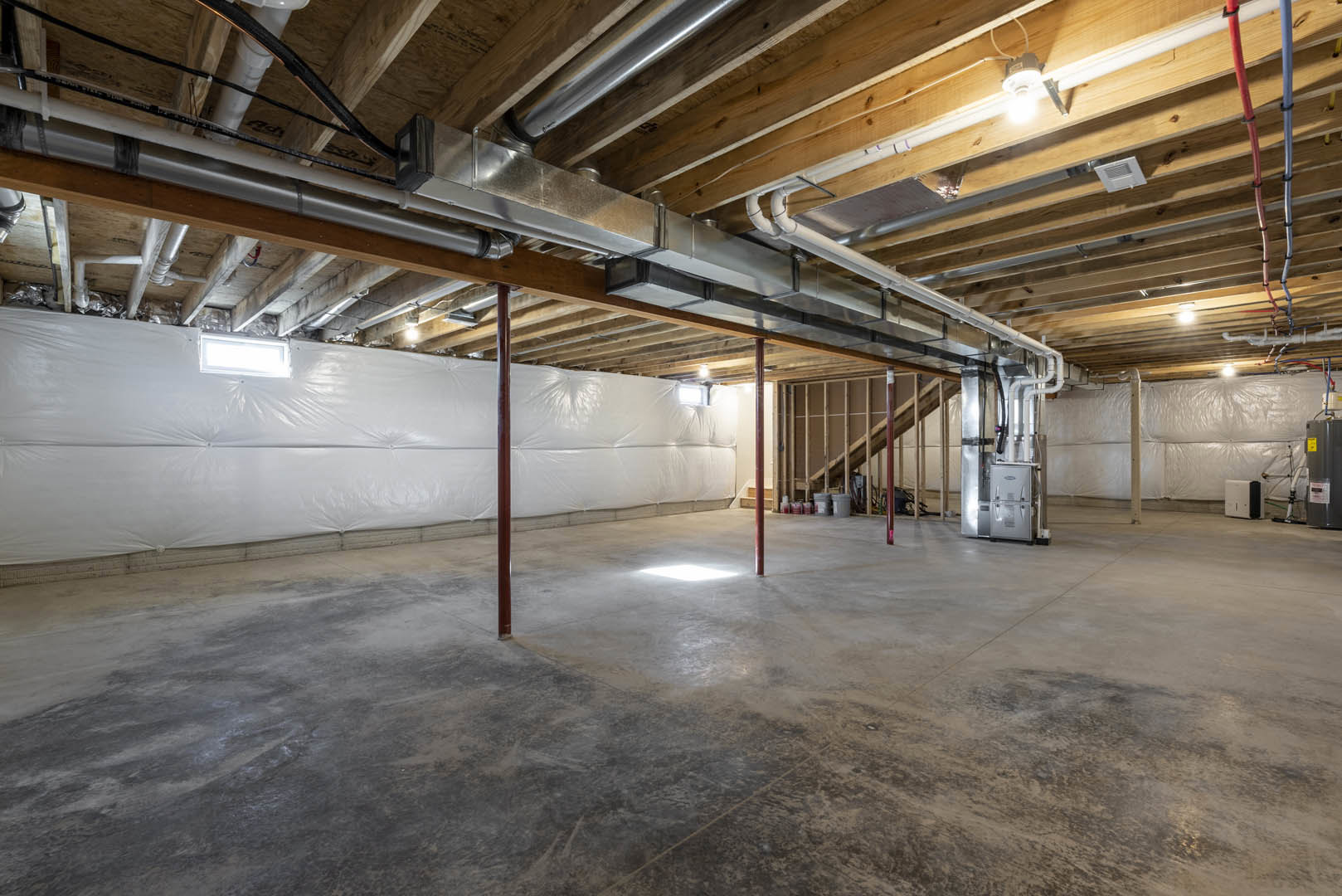 Exposed metal pipes and beams in unfinished basement with concrete floor, red support poles, white vented object, plastic-covered wall, and ceiling light fixture