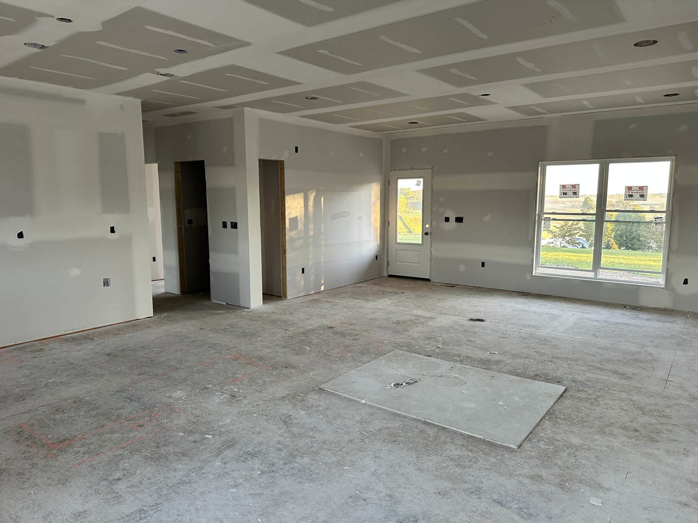 Concrete-floored room with large windows, glass-paneled door, plaster walls, and ceiling; visible broken tile and hole in floor, lamp fixture near entry.
