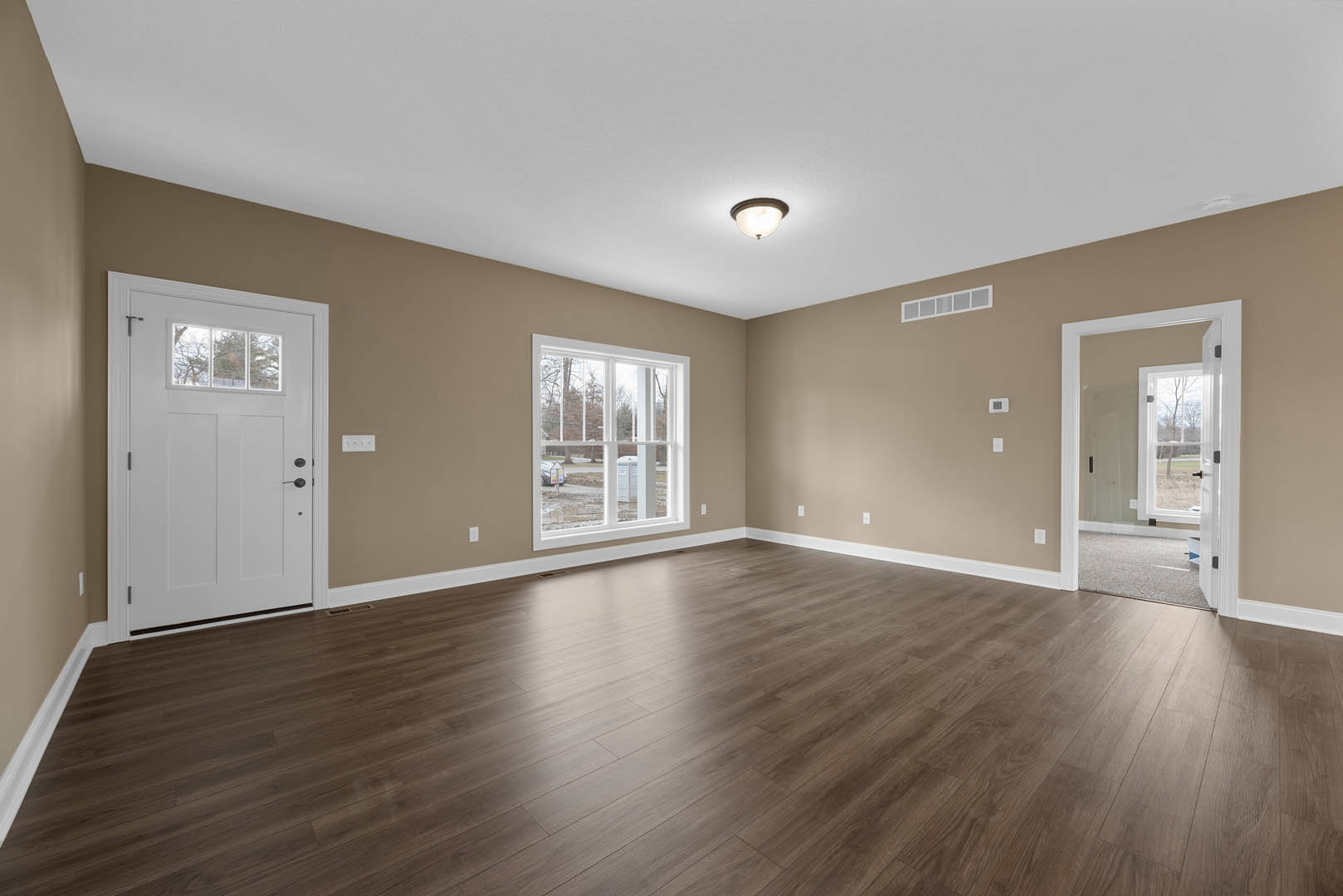 Room with light wood flooring, white plaster walls, white door featuring a glass window, ceiling-mounted light fixture, and window overlooking parking lot and landscape.