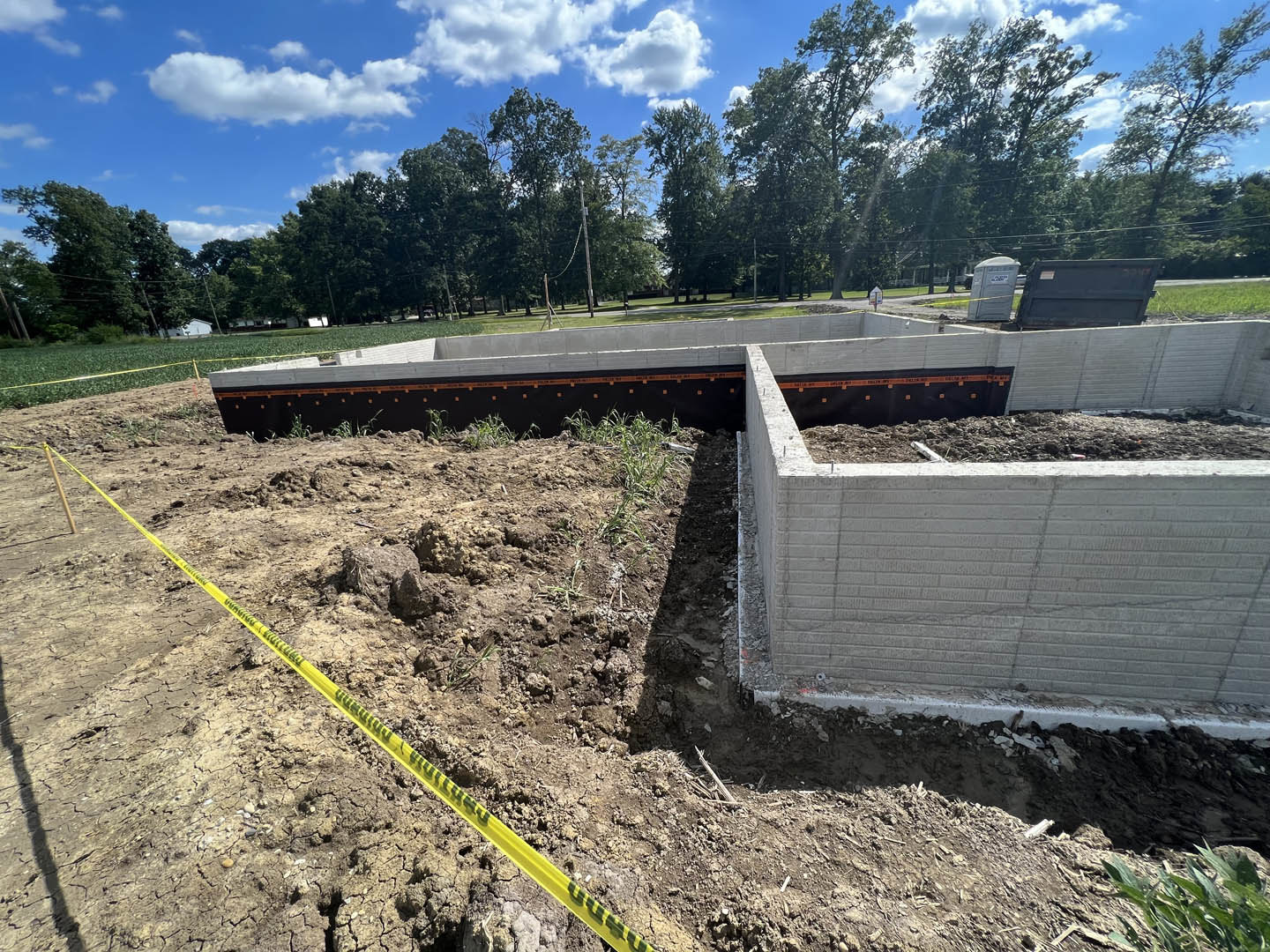 Concrete foundation surrounded by dirt and construction tape, partially built wall with exposed hole, portable toilet on street, man standing near wall, trees and blue sky in