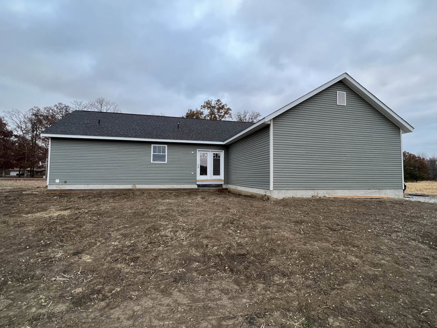 Grey siding house with white double doors, glass windows, small porch, and dirt yard under cloudy sky