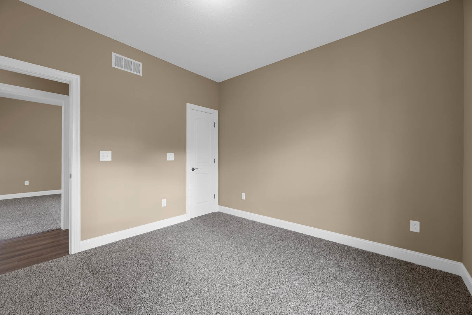 Beige-walled room with white paneled door, carpeted floor, wall vent, and white trim molding