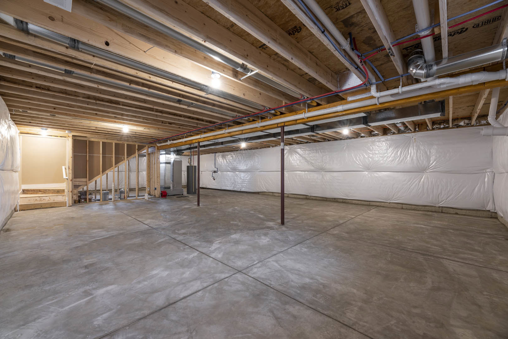 Basement room with exposed black and white pipes along white tile and drywall, grey concrete floor, wooden beam ceiling, and white plastic wall covering.