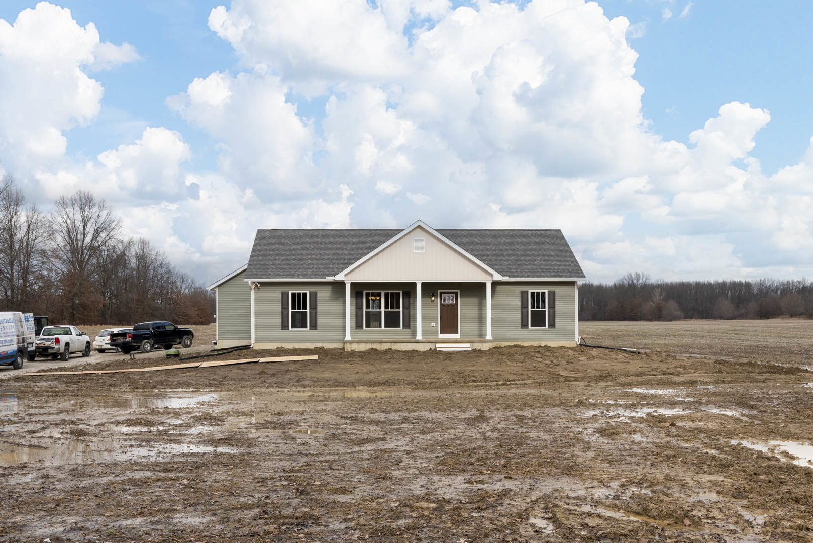 Two-story custom home with grey roof, white siding, and large windows; muddy ground in front, black and white trucks parked nearby, cloudy sky overhead.