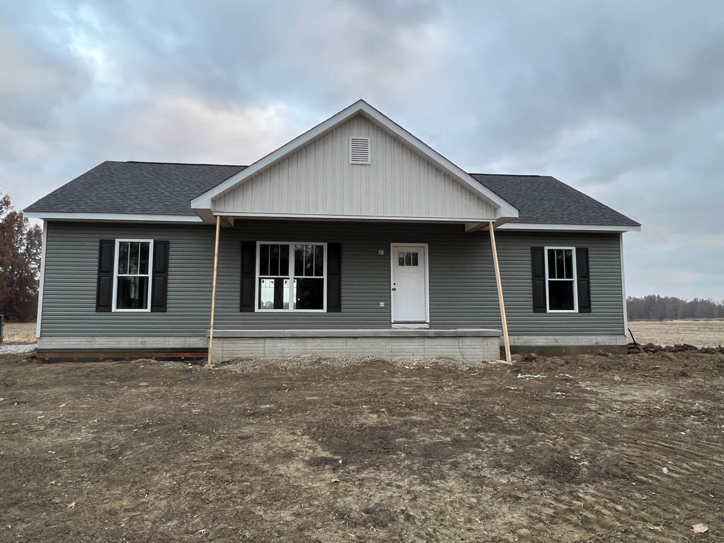 Partially built house with white siding, black-framed windows, and white door, surrounded by dirt lot under cloudy sky