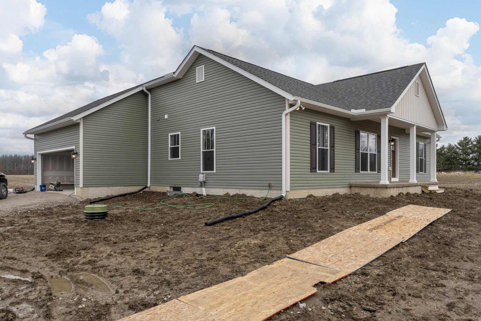 Grey siding house with large windows, plywood sheets on dirt ground, green-lidded black container near a path, cloudy sky overhead.