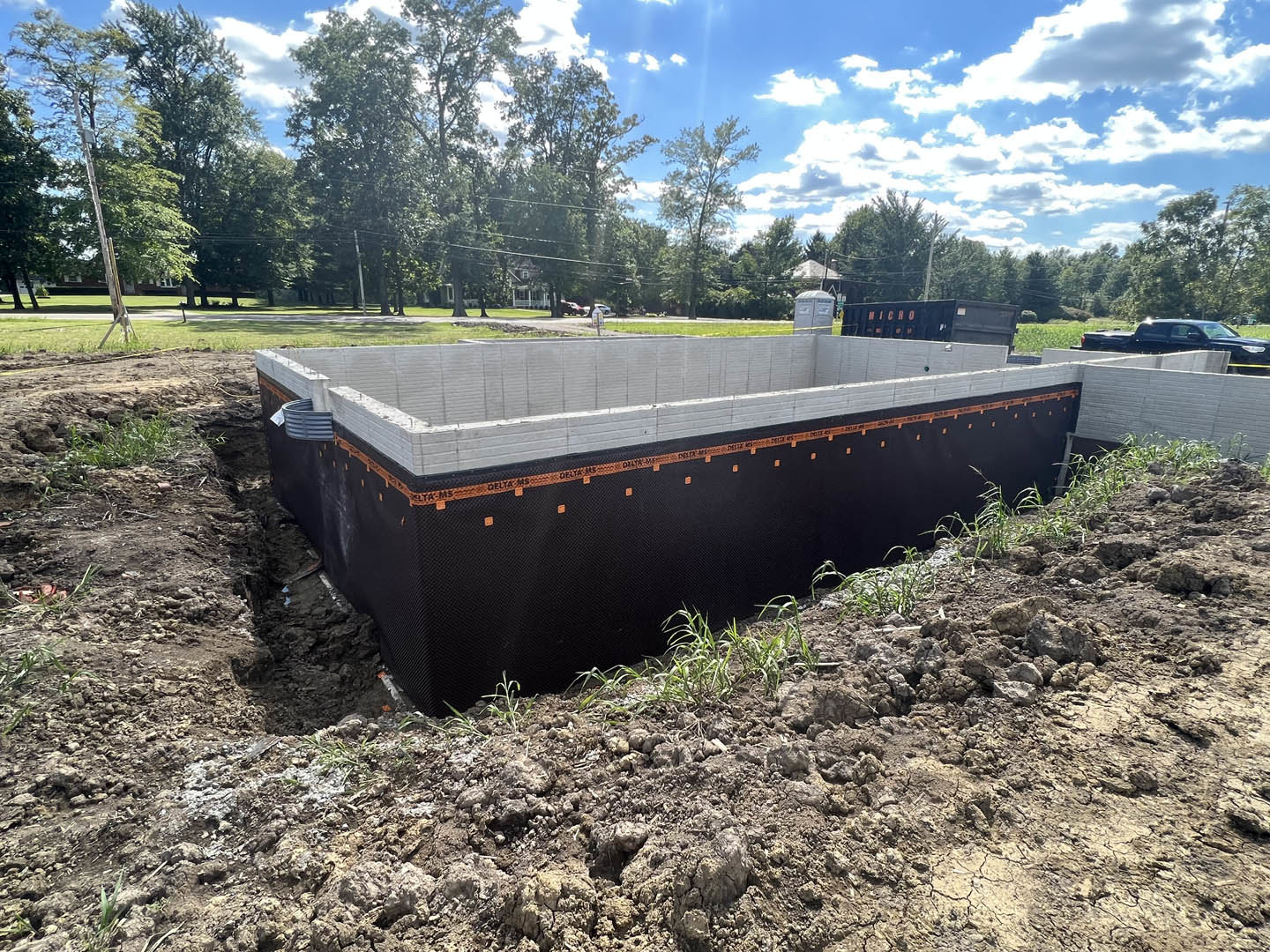 Concrete house foundation with orange tape along black and white perimeter wall, blue sky overhead, trees in background, grey portable toilet on grassy area, parked car on nearby