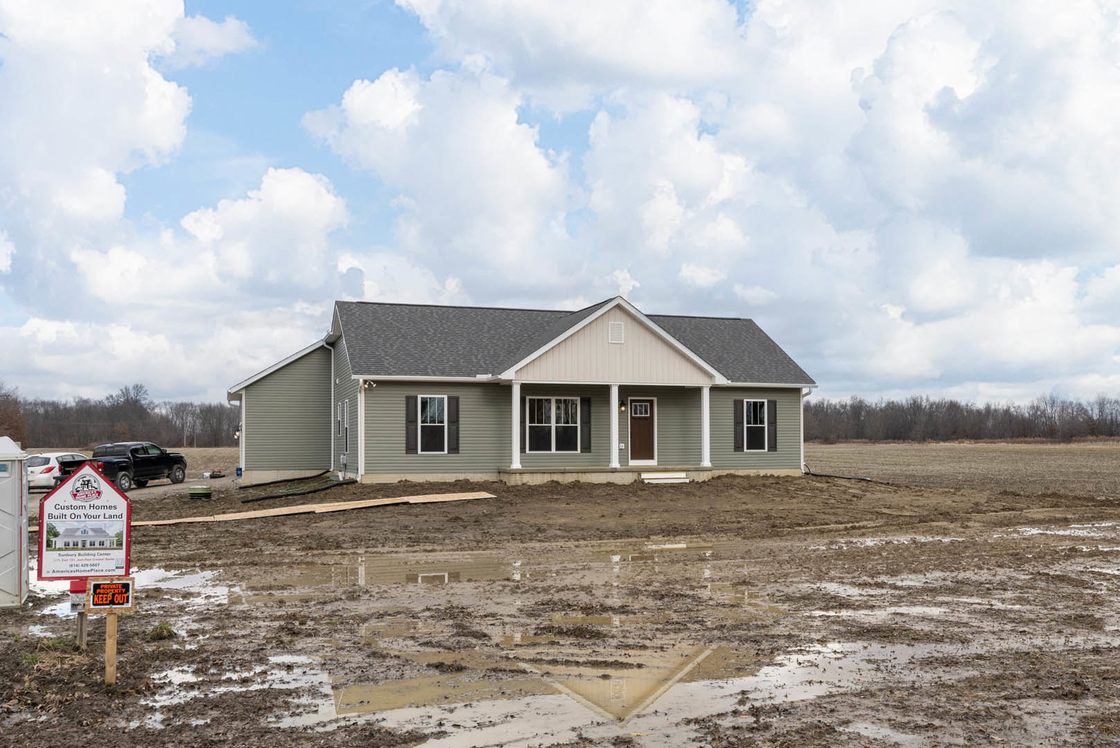 Two-story house with grey roof, white trim double doors, several windows, muddy ground with puddles, construction sign displaying house illustration, overcast sky, sparse trees in