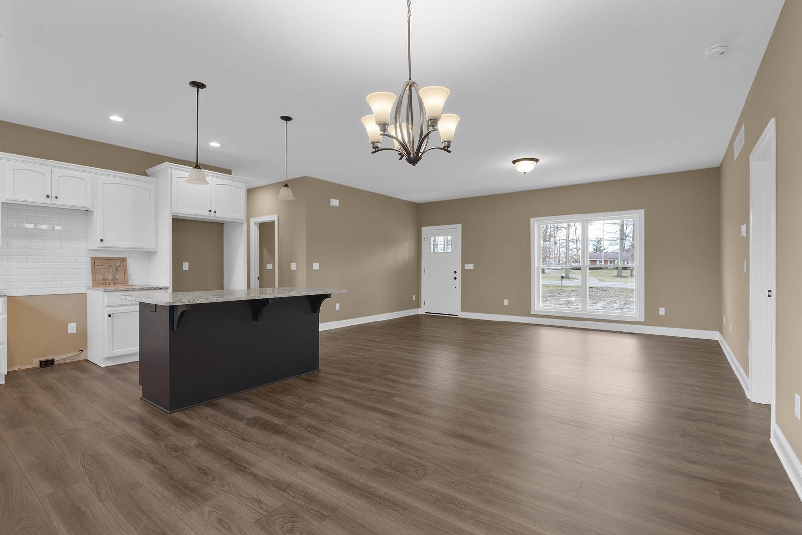 Spacious kitchen with black marble-topped island, wood flooring, white cabinetry, large window overlooking street and trees, white door with glass panel, modern chandelier