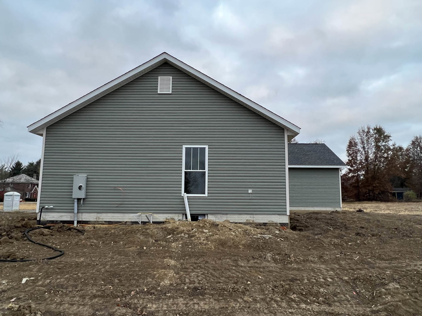 Grey house with white-framed window and white vent, large dirt patch in foreground, metal pipe protruding from dirt hill, white plastic container near foundation, cloudy sky above.