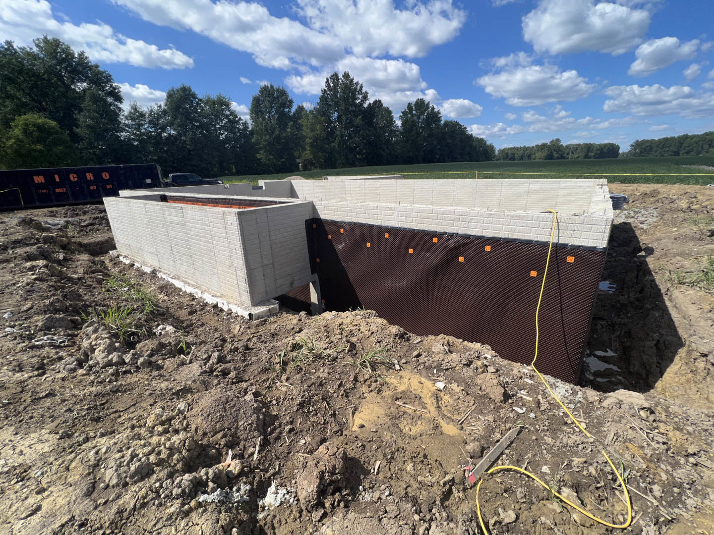 Concrete foundation and framed walls surrounded by dirt and construction fencing, with trees and blue sky in the background