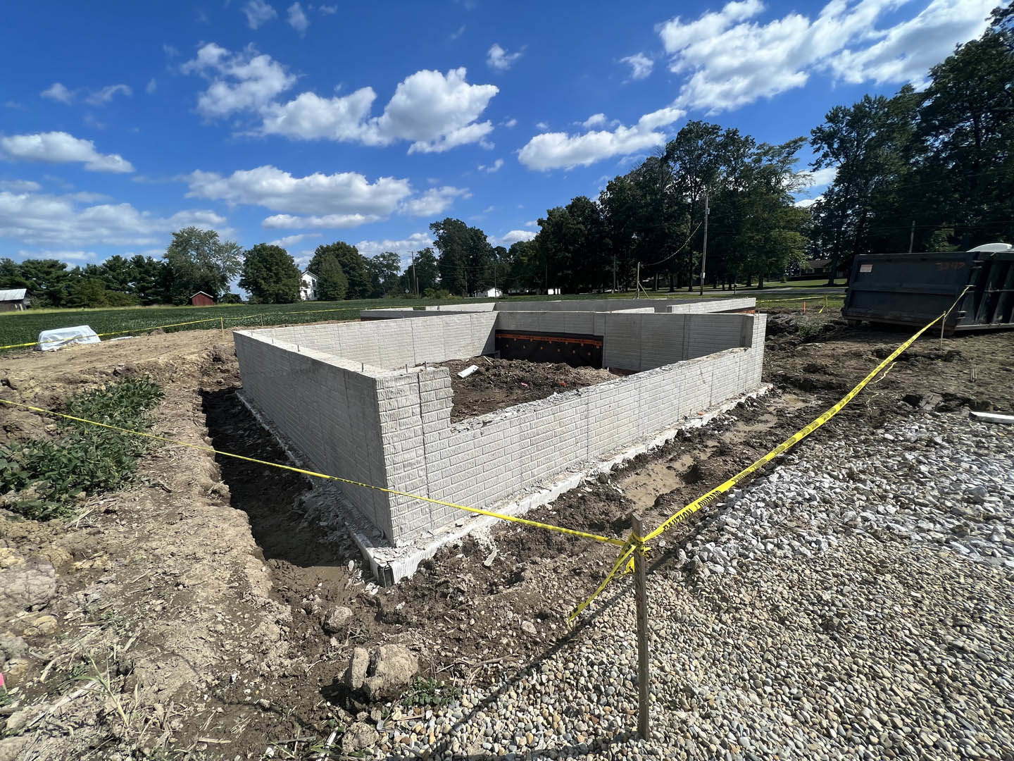 Partially built brick wall on a residential construction site, surrounded by dirt piles, yellow caution tape, and a dump truck, with trees and clouds under a blue sky