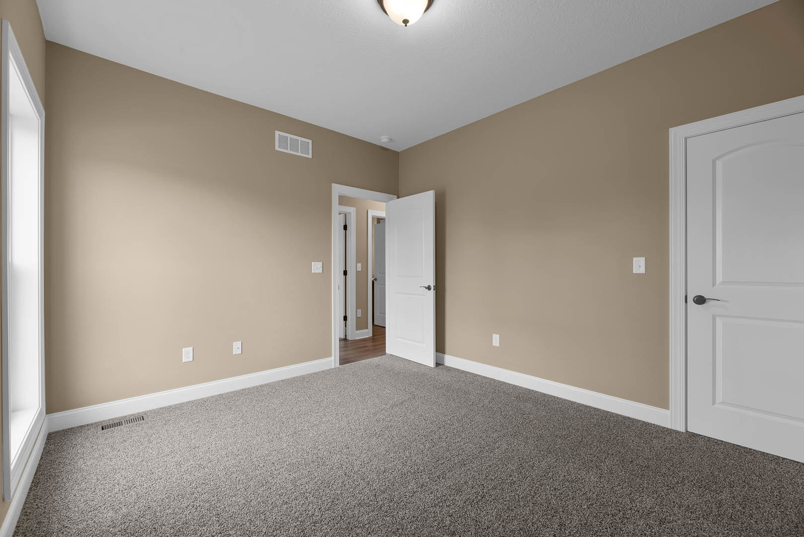 Beige-painted room with white rectangular door featuring silver handle, carpeted floor, wall vent, and light streaming through open doorway