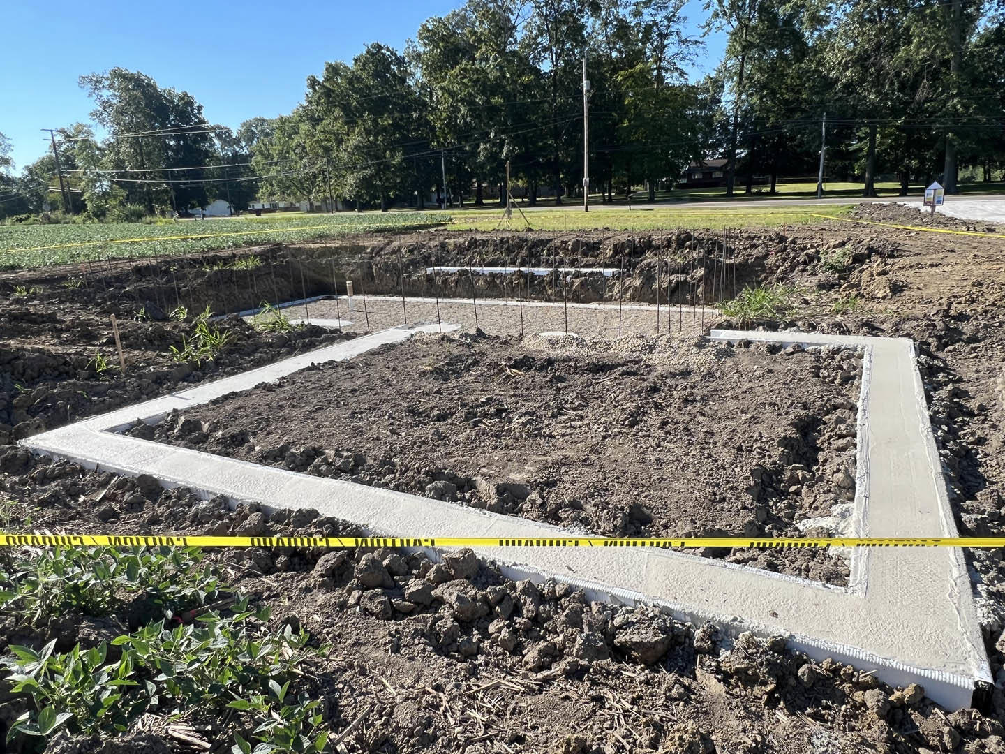 Construction site with exposed soil, rocks, and green plants, yellow caution tape stretched across foreground, leafy tree in background