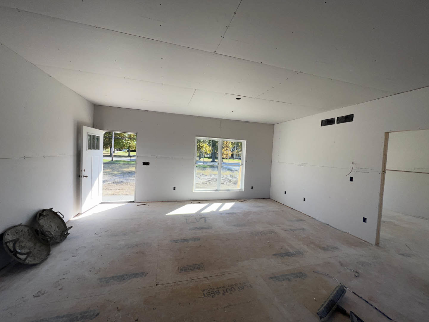 Spacious room with white plaster walls, unfinished ceiling with visible holes, large window overlooking yard and trees, and door leading outside