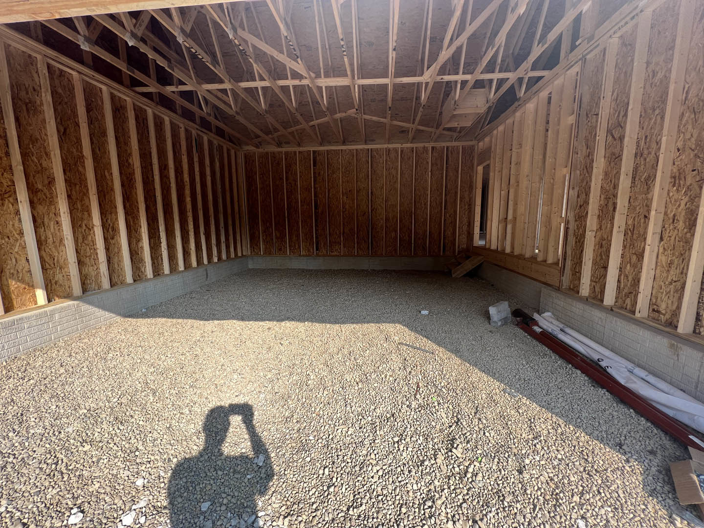 Person’s shadow cast on unfinished drywall in a room under construction, exposed wooden beams overhead, stack of grey bricks and gravel ground visible