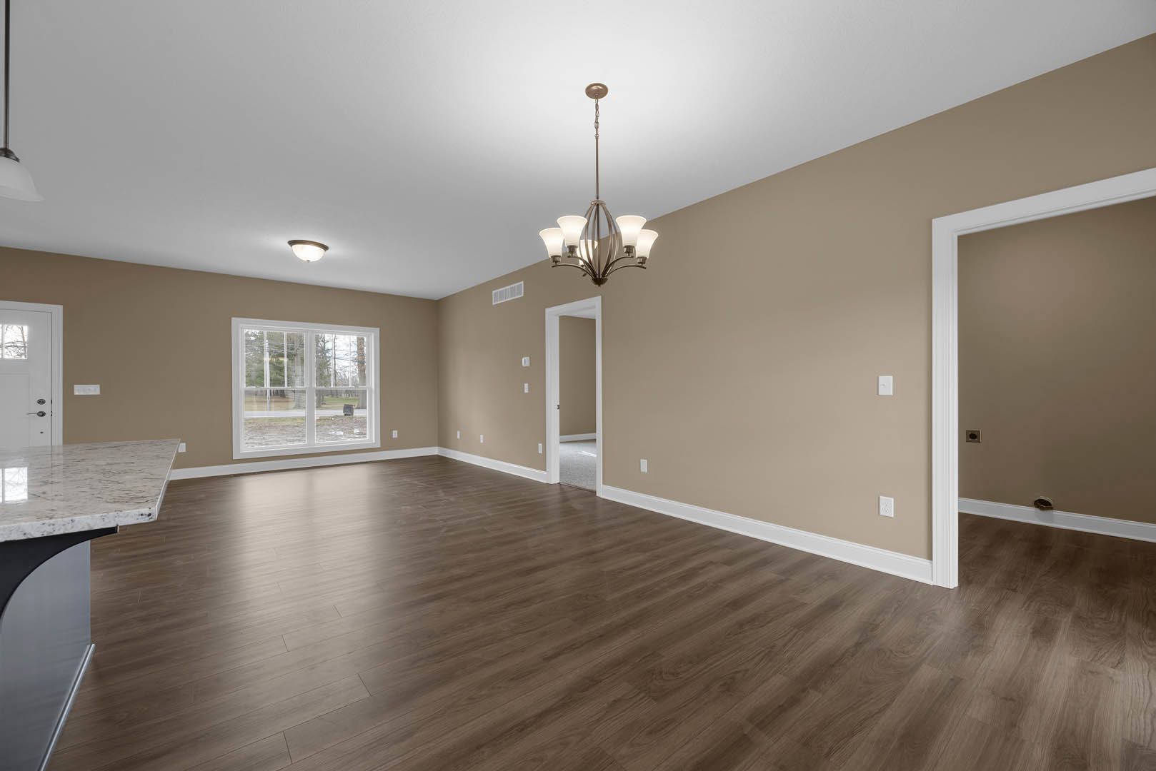Open living space with hardwood flooring, white baseboards, large window overlooking road, ornate chandelier hanging from plaster ceiling, neutral walls.
