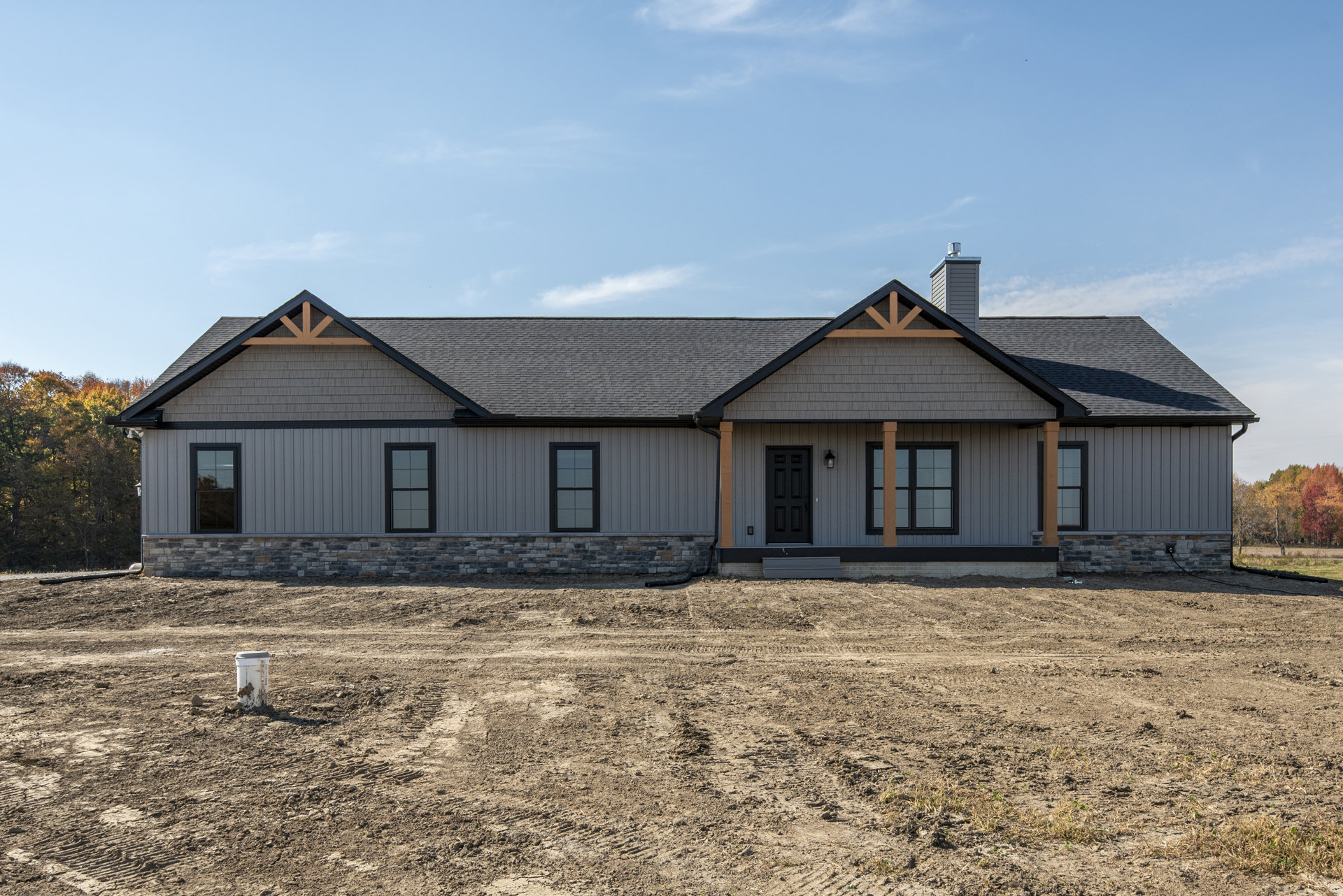 Modern house with black front door, large windows, and light-colored siding, set on a dirt field with visible tire tracks under a cloudy sky