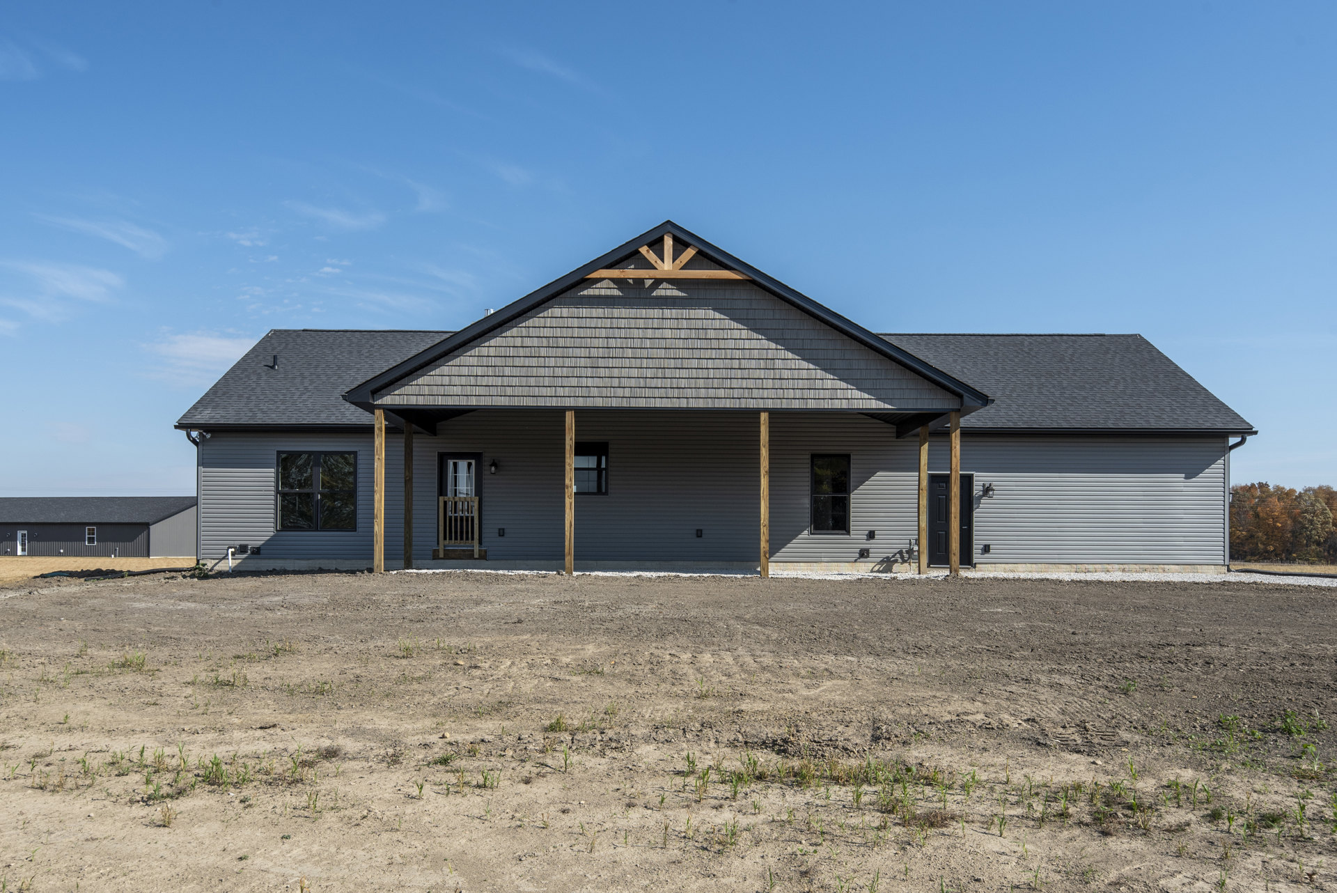 Modern house with black-framed windows, wooden railing, and pitched roof, surrounded by a large dirt field and fenced boundary under a partly cloudy sky