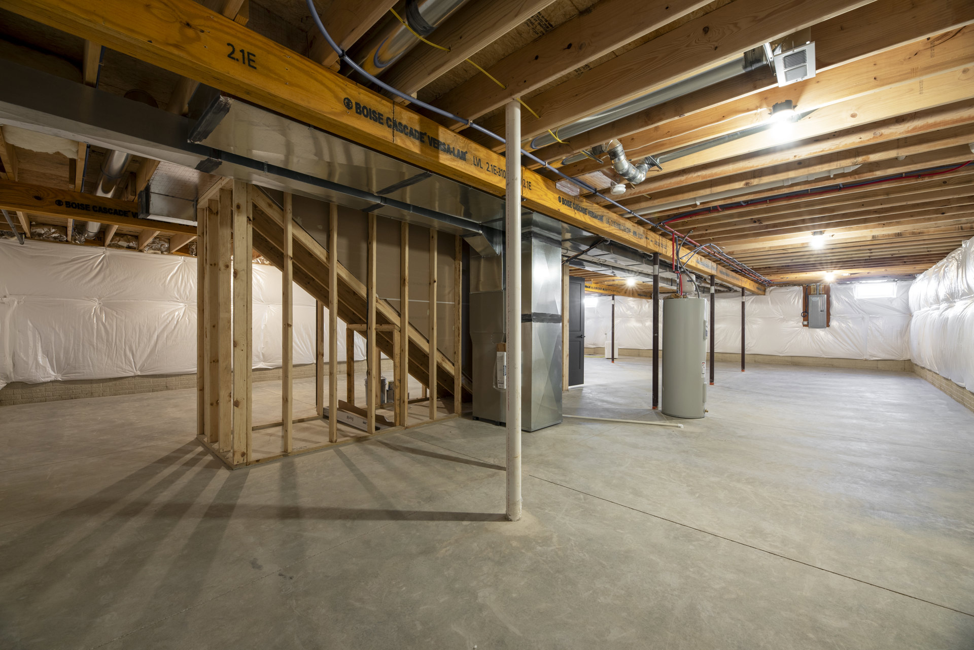 Open room with exposed wood ceiling beams, steel pipes, and a staircase; white cylindrical appliance with open door sits near unfinished walls and concrete floor.