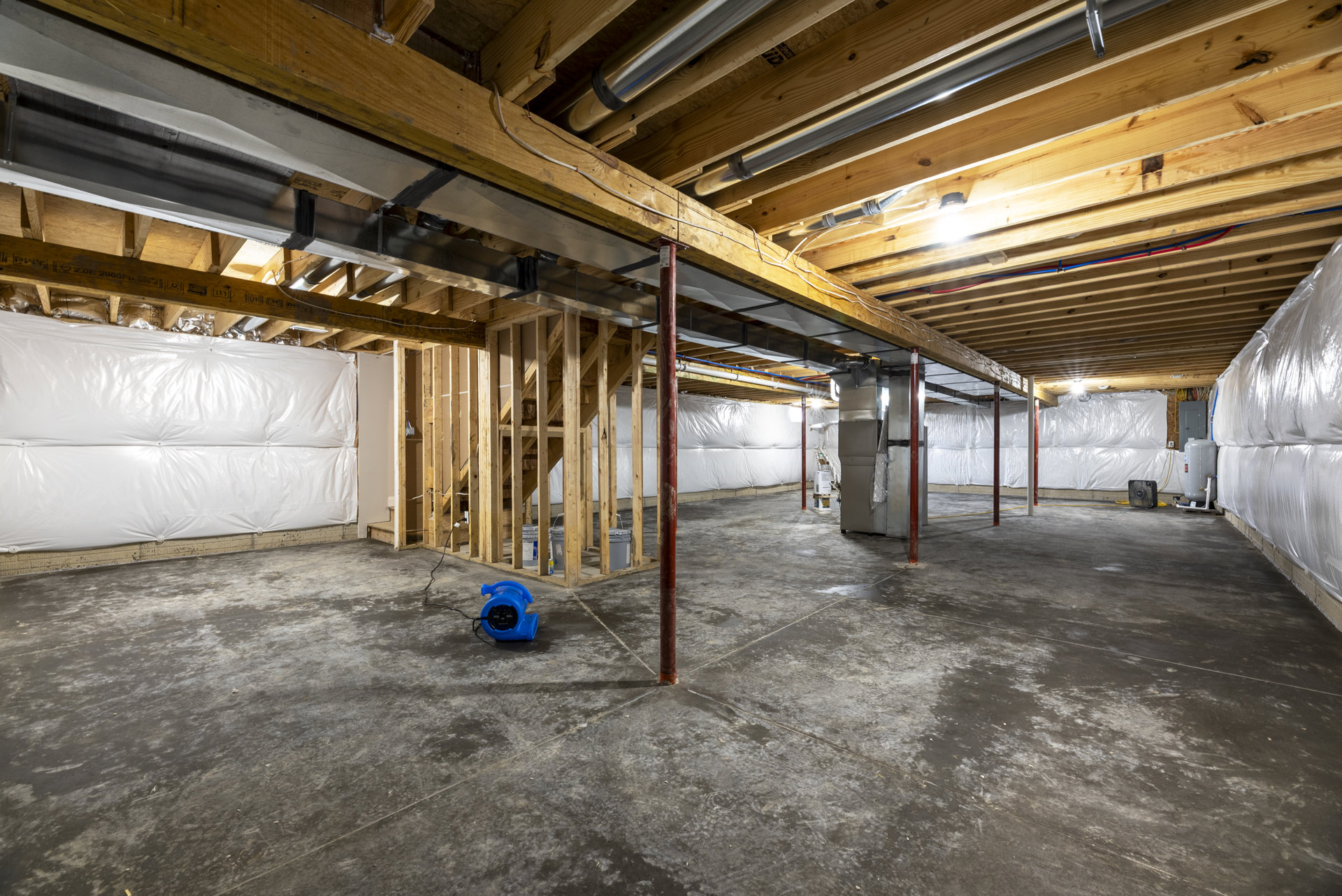 Concrete basement room with exposed wooden ceiling beams, blue hose and pipe, blue blower fan, red pole, white plastic bag, and metal pipes visible.