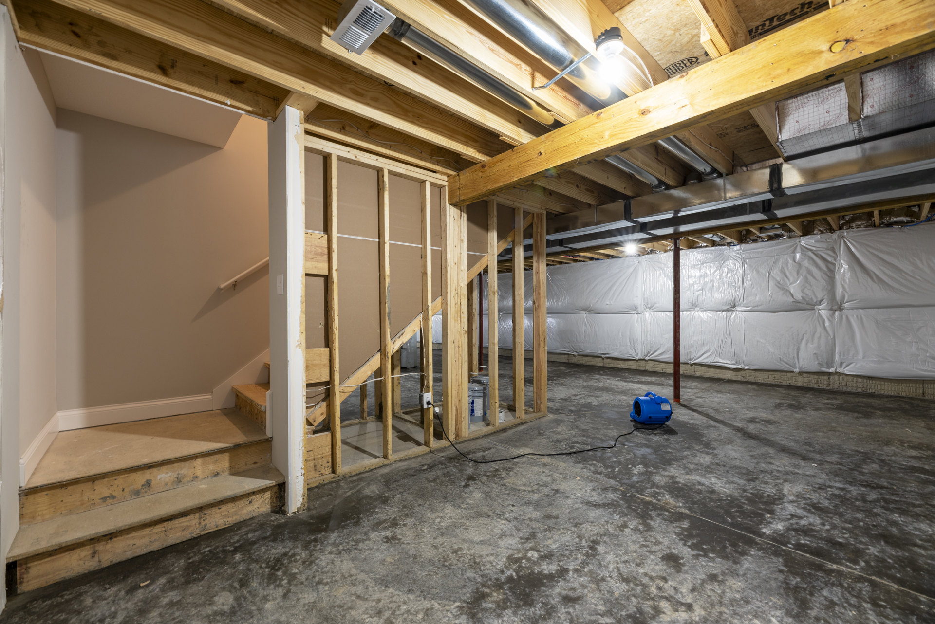 Unfinished basement with exposed wood beams, white plastic wall covering, blue machine on concrete floor, electrical wire plugged into wall