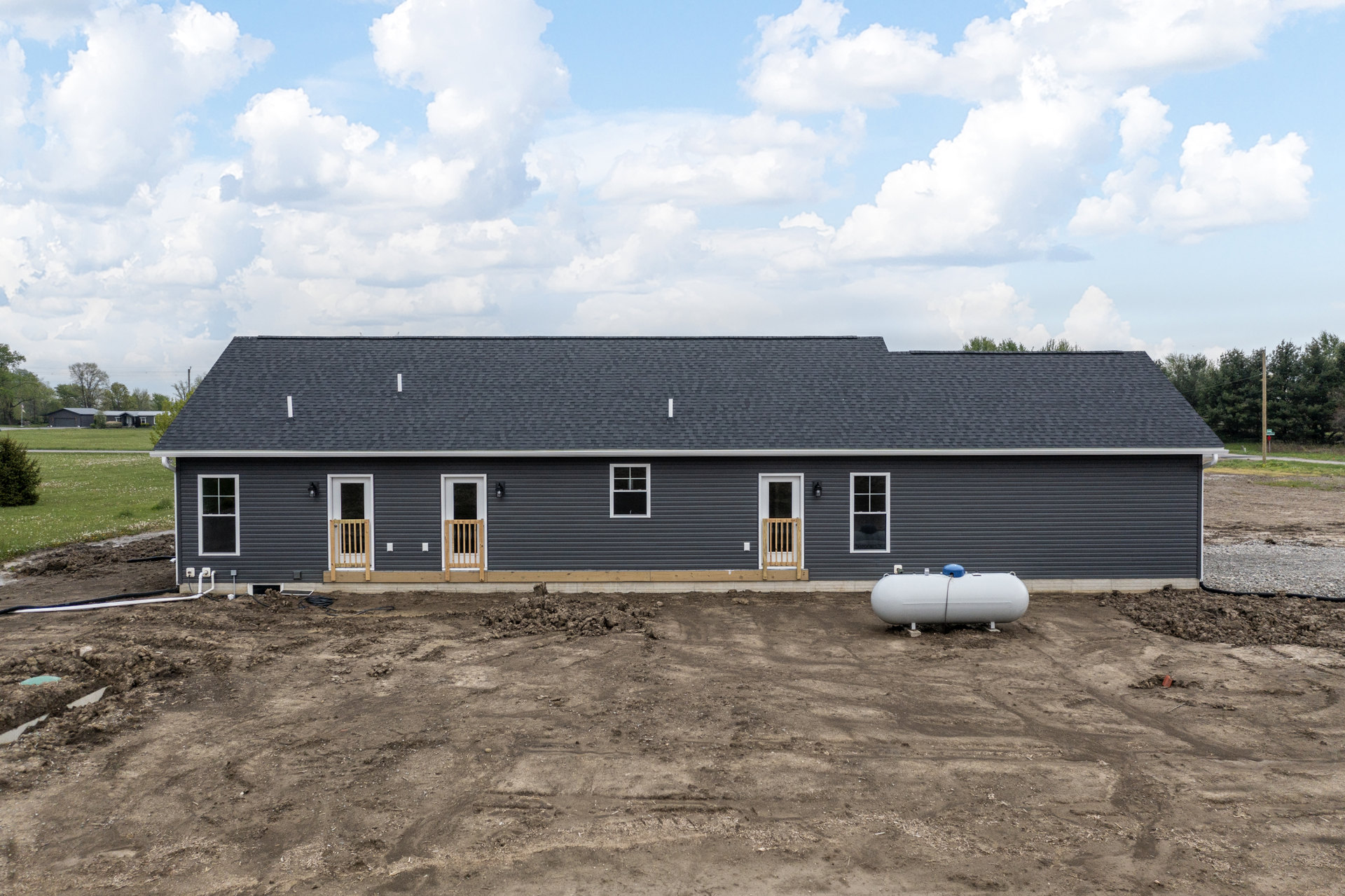 White-framed window and open door on house exterior, large cylindrical tank partially buried in dirt, wooden fence section near entry, cloudy sky above roof, sparse grass and trees