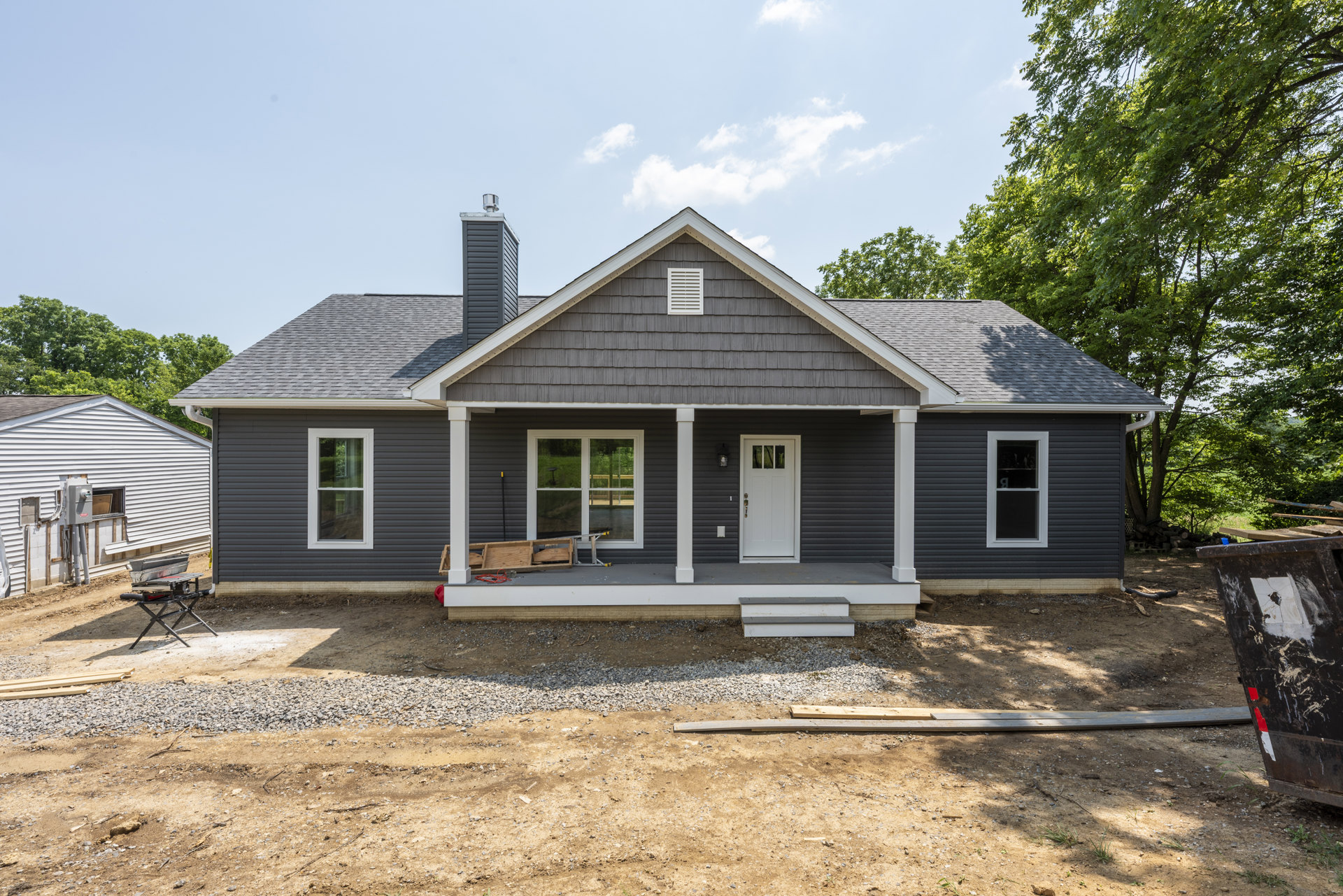 Gray house with covered porch, gravel driveway, white vent on exterior wall, white door with window, table holding a saw, black metal object with white and red tape, small white