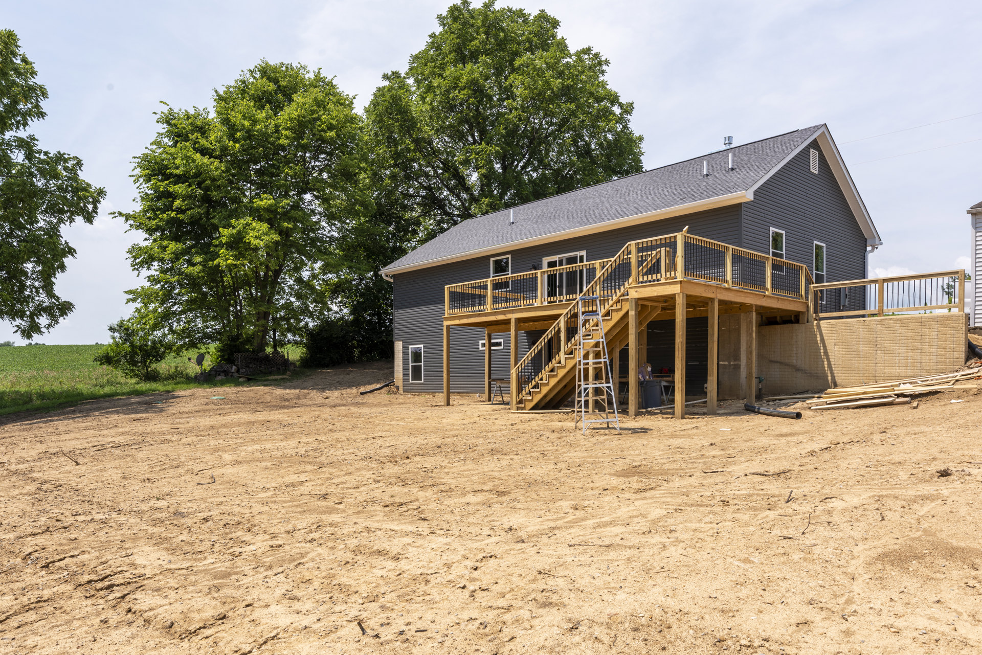 Wooden deck with stairs and ladder attached to a house, overlooking a dirt field with a leafy tree nearby