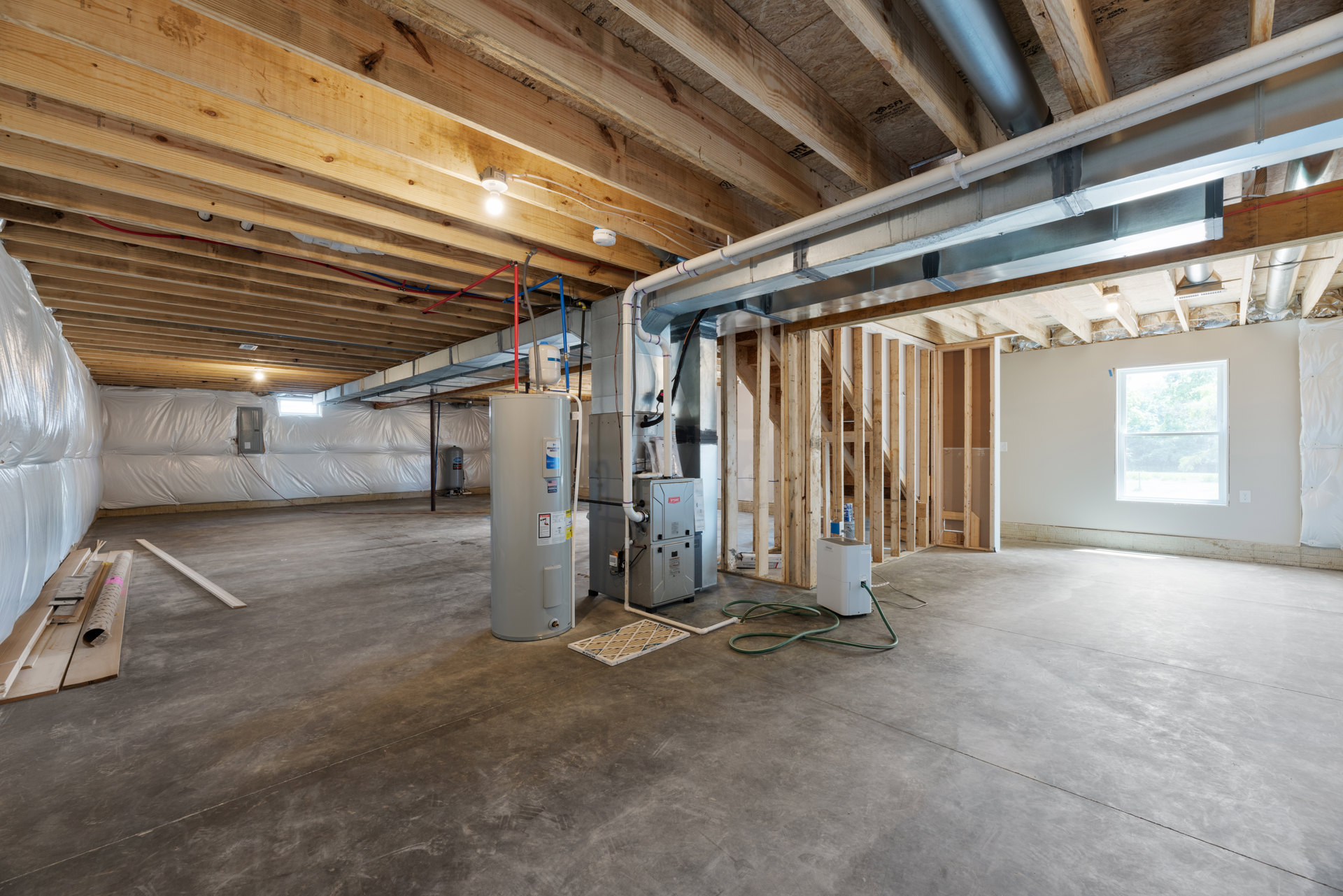 Basement utility room with exposed metal beams, large white cylindrical water heaters with blue labels, visible pipes and filter, window overlooking trees, unfinished walls and