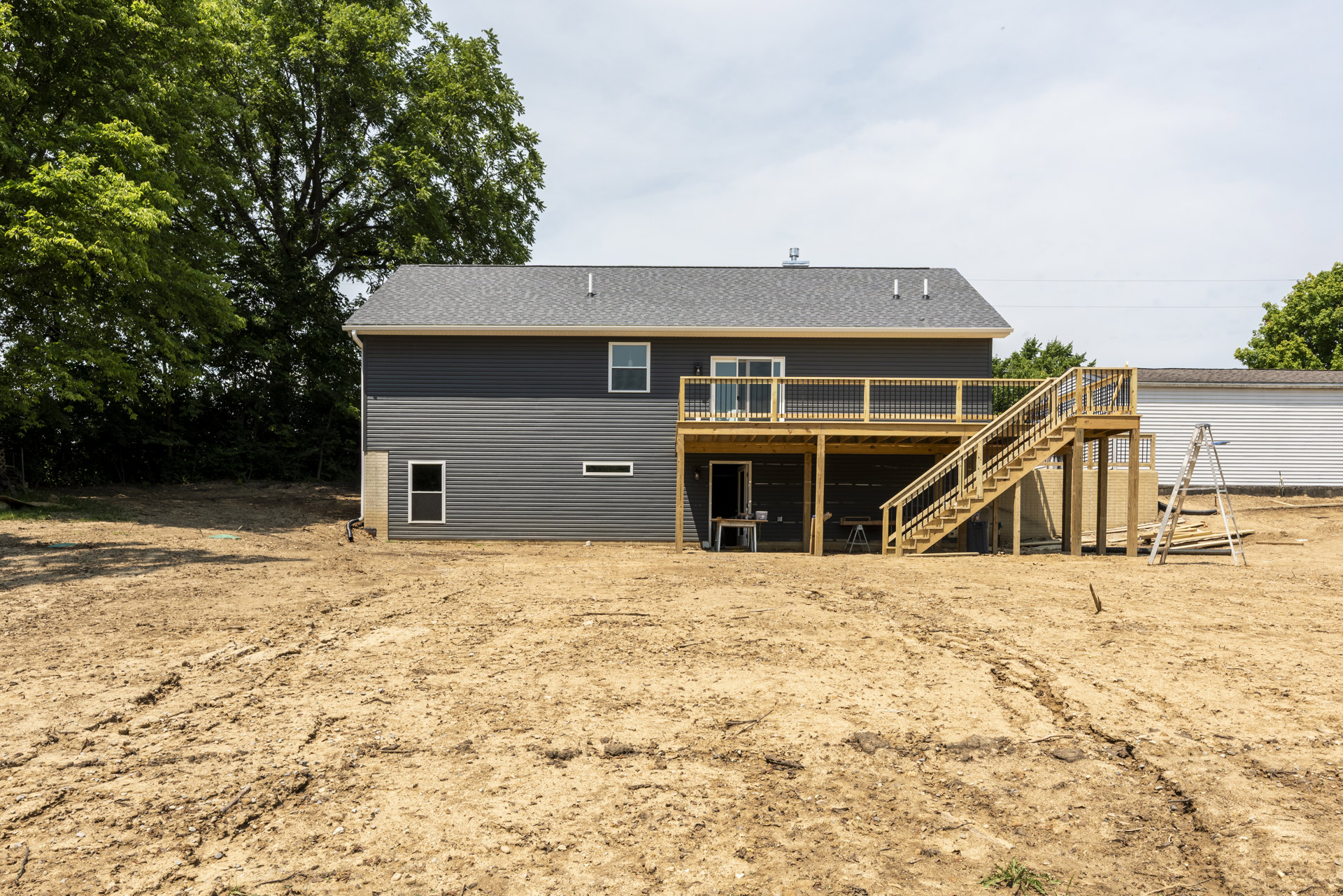 Two-story house with light siding, white-framed windows, elevated wooden deck, fenced yard, and mature tree in the background.
