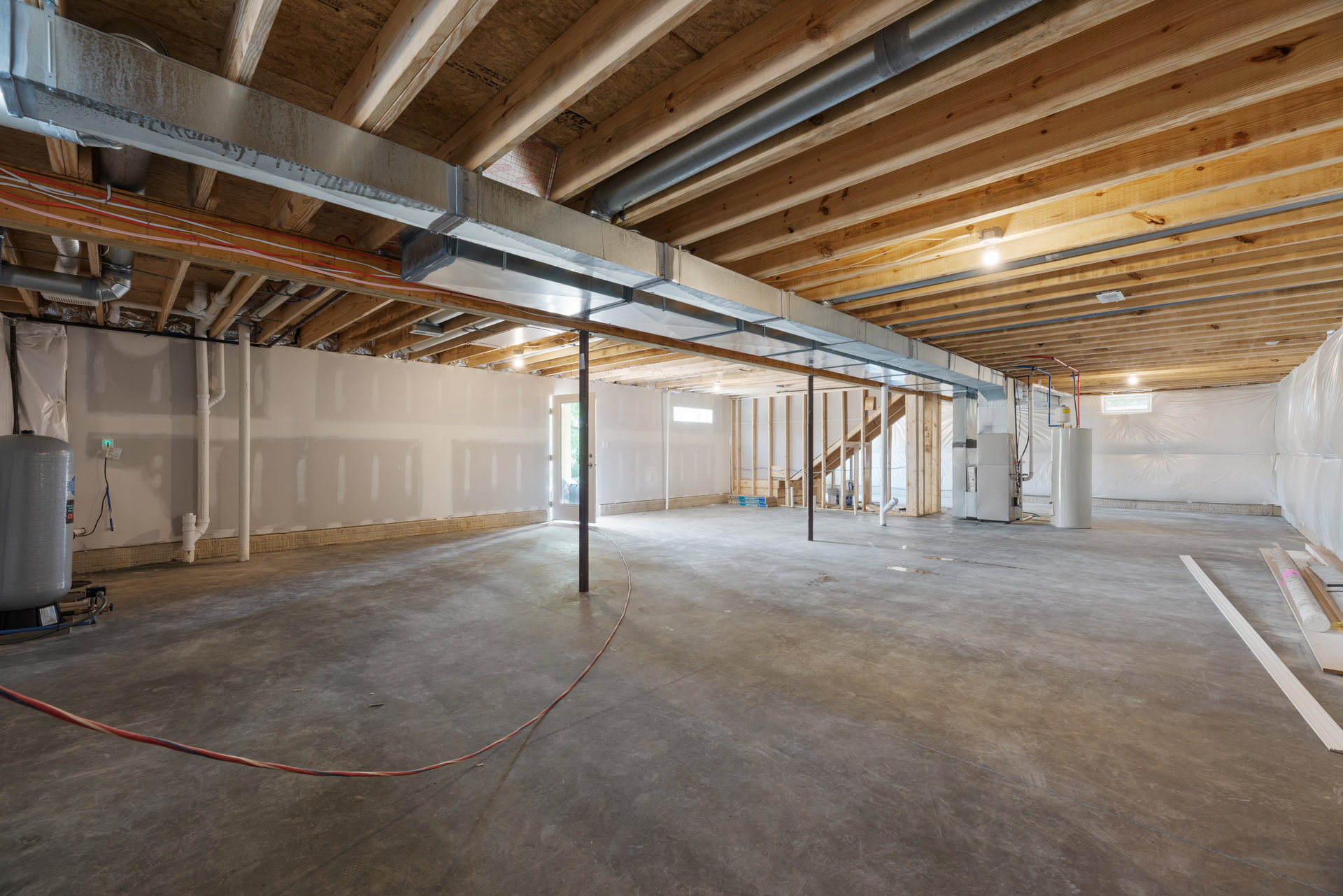 Exposed wooden ceiling beams above a concrete floor, visible metal pipe and red and white electrical cables, unfinished walls with insulation in a basement interior